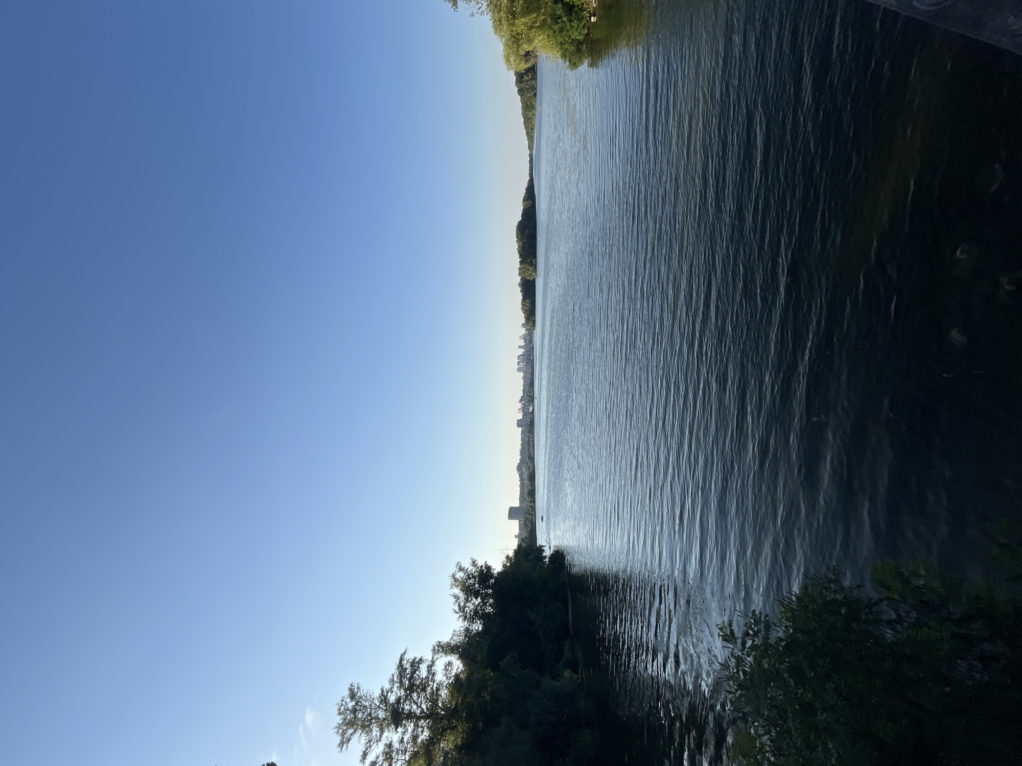 This image shows a wide river with rippling water, reflecting the clear blue sky. On the left bank, a dense line of dark green trees and foliage extends into the foreground. The right bank is also lined with green trees. In the distance, across the river, a cityscape with buildings and a few scattered trees can be seen under a bright sky. There are small boats visible on the water in the distance, and possibly a few people on the bank. The overall scene suggests a calm, sunny day in an urban environment with significant natural elements. The vantage point appears to be from a bridge or elevated area overlooking the river. This scene is likely the Dnieper River in Dnipro, Ukraine, given its characteristic wide expanse and the distant cityscape.