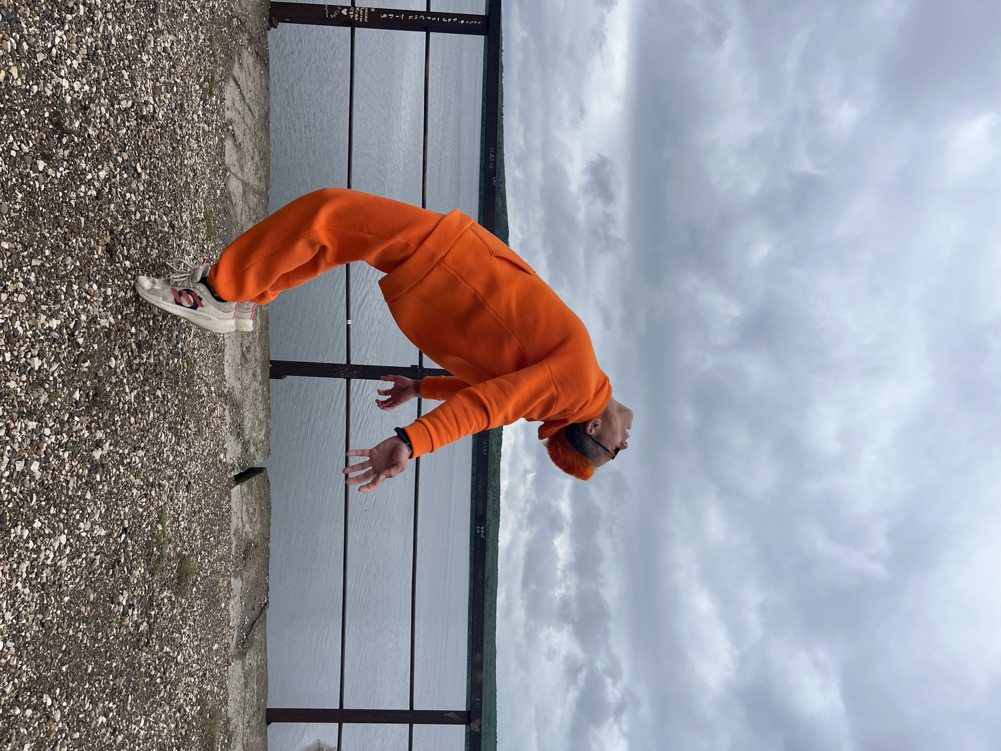 A man wearing an orange hooded sweatshirt and sweatpants, standing on a pebble-covered embankment, is bent backward with his hands outstretched. He is wearing sneakers and sunglasses. Behind him is a metal railing, a body of water, and trees in the distance under a cloudy sky. The location is Chaikovsky, Russia.