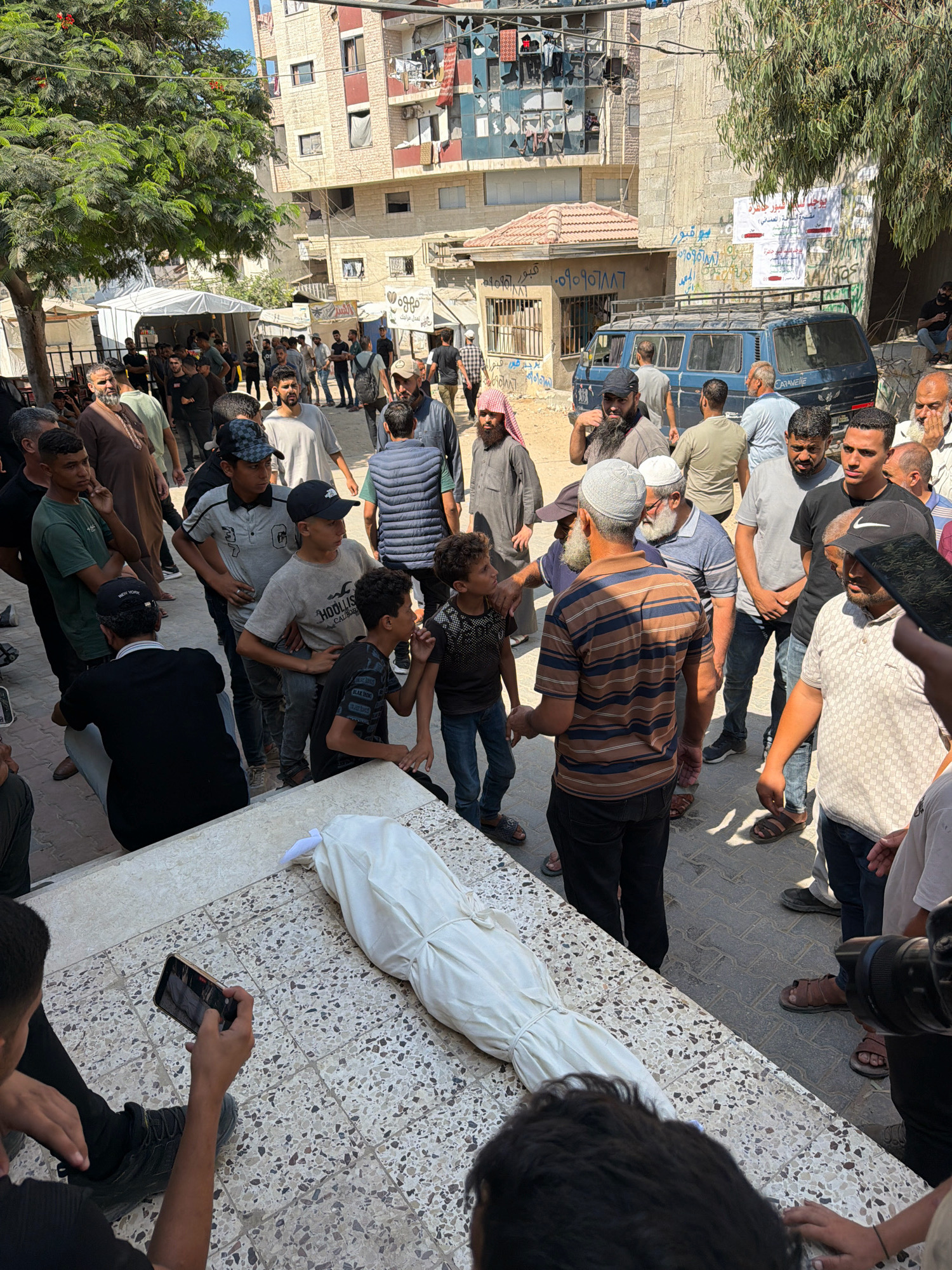 In a street in Gaza City, Palestinian Territories, a group of men are gathered around a body covered in a white shroud. The body is laid out on a tiled surface. Some men are looking at the body, while others are talking or observing. One person is holding up a phone, appearing to record the scene. The environment is urban, with buildings visible in the background, including a blue van and what appears to be a market tent. The overall atmosphere is somber.