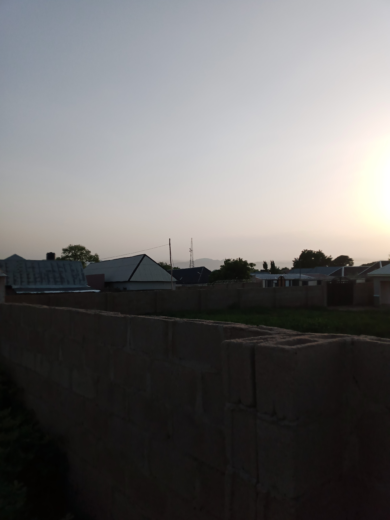 This media file depicts an outdoor scene at dusk in Jauro Yinu, Nigeria. The foreground is dominated by a wall constructed from large, hollow concrete blocks, casting a dark shadow. Beyond the wall, several buildings with corrugated metal roofs are visible, suggesting a residential or semi-urban area. A tall telecommunications tower stands prominently in the mid-ground, hinting at infrastructure development. Trees are scattered throughout the landscape, some appearing lush and green, indicating a tropical or subtropical climate. The sky is a pale gradient, transitioning from a light gray at the top to a soft, yellowish hue near the horizon where the sun is setting. The overall atmosphere is peaceful and quiet, characteristic of the end of a day. There are no people or animals visible in the frame. The lighting suggests it is either early morning or late evening. The presence of the concrete block wall and the style of the buildings could be indicative of a developing area.