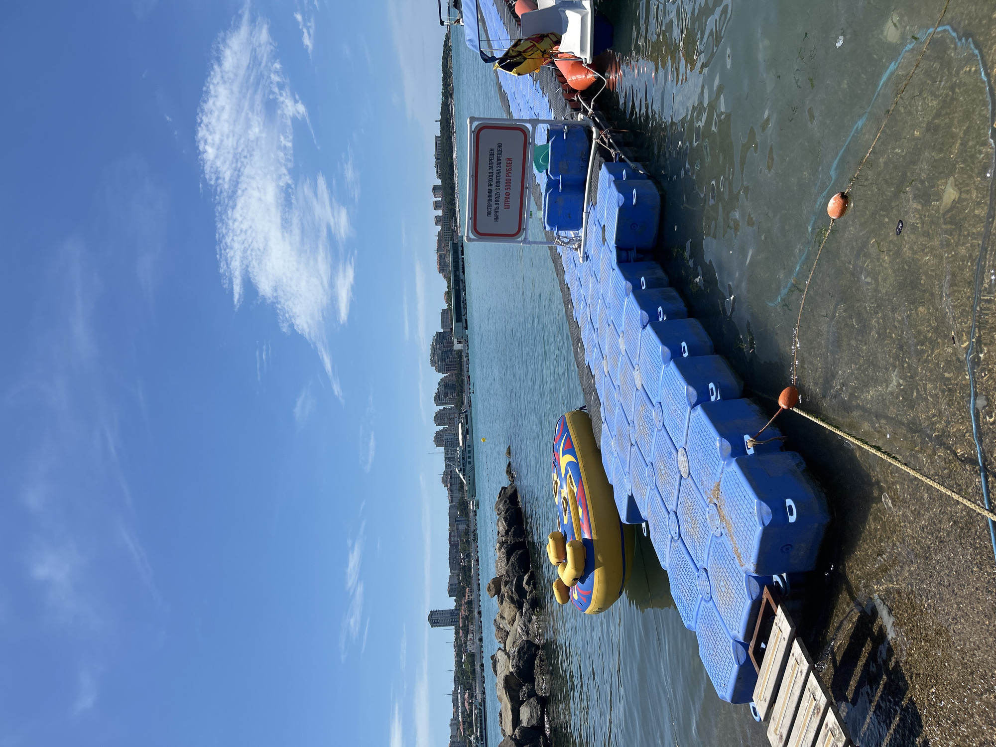 A sunny day scene at the Gelendzhik embankment in Russia, featuring a bright blue sky with sparse white clouds. In the foreground, a modular blue plastic pontoon dock extends into the calm, clear water. A yellow and blue inflatable tube, likely for recreational use, floats tethered to the dock. To the right of the dock, a small white boat with orange fenders is partially visible. A sign on the dock, written in Russian, warns against jumping into the water from the pontoon and states a fine of 5000 rubles. In the background, a cityscape with buildings and a large white ferry can be seen across the bay, with a breakwater made of large rocks in the middle ground. The water is a deep blue, reflecting the sky.