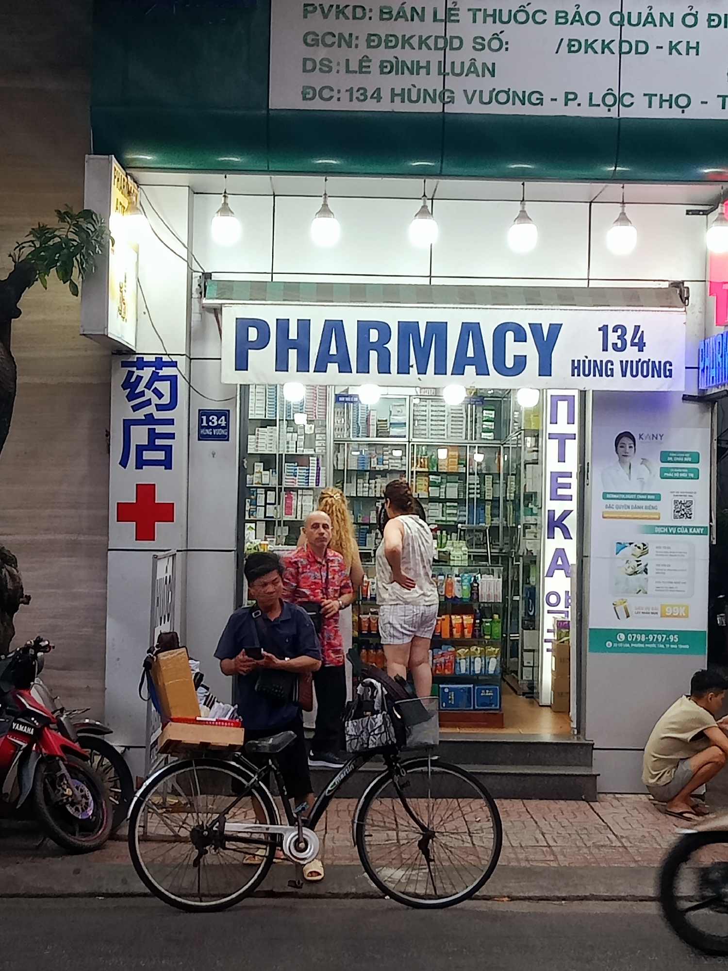 This media file captures a nighttime street scene in Nha Trang, Vietnam, centered around a brightly lit pharmacy. The storefront is predominantly white, featuring the prominent blue text "PHARMACY" and the address "134 HÙNG VƯƠNG". To the left, a vertical sign displays Chinese characters "药店+" (Pharmacy) and a red cross, with "134 HÙNG VƯƠNG" written below. Above the main sign, Vietnamese text provides pharmacy details, including "PVKD: BÁN LẺ THUỐC BẢO QUẢN Ở ĐI" (Retail sale of medicines stored at...), "DS: LÊ ĐÌNH LUÂN" (Pharmacist: Le Dinh Luan), and the full address "ĐC: 134 HÙNG VƯƠNG - P. LỘC THỌ - T" (Loc Tho Ward, Nha Trang City).
Inside the pharmacy, shelves stocked with various products are visible. Several people are present: a man with a bald head and a red patterned shirt stands near the entrance, looking out, with a woman with long, light-colored hair partially obscured behind him. Another woman, wearing light-colored shorts and a striped tank top, stands deeper inside, facing the shelves.
Outside, in the foreground, a man in a dark blue shirt and shorts sits on a black bicycle, looking at his phone. The bicycle's rear rack holds a large brown box and a small basket. A red and black Yamaha motorbike is parked to the left. On the right side of the frame, another man in a light t-shirt and shorts squats on the sidewalk. A vertical sign on the right side of the pharmacy displays "ПТЕКА" (likely part of "APTEKA" or a stylized equivalent) and an advertisement for "KANY" featuring a woman's face, details on "DERMATOLOGIST CHAU DUU", and a phone number "0798-9797-95". The scene depicts a typical evening at a local pharmacy, with individuals seemingly either waiting, browsing, or working.