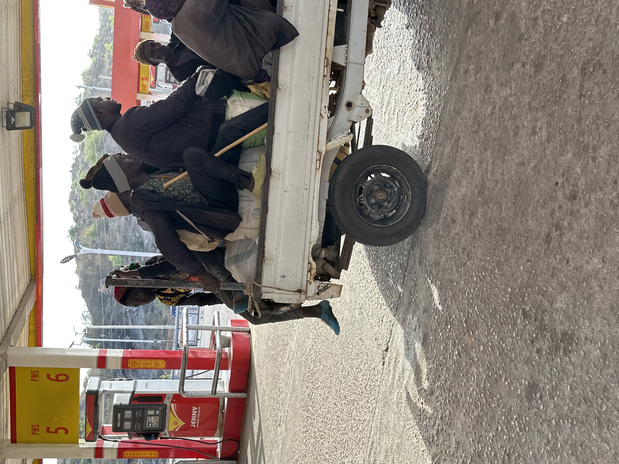The image shows a white truck at a Jenny fuel station in Jos, Nigeria. The pump is yellow and red, marked with "PMS 5" and "PMS 6." People are riding in the back of the truck, including one wearing blue shoes. The background shows a hilly landscape.