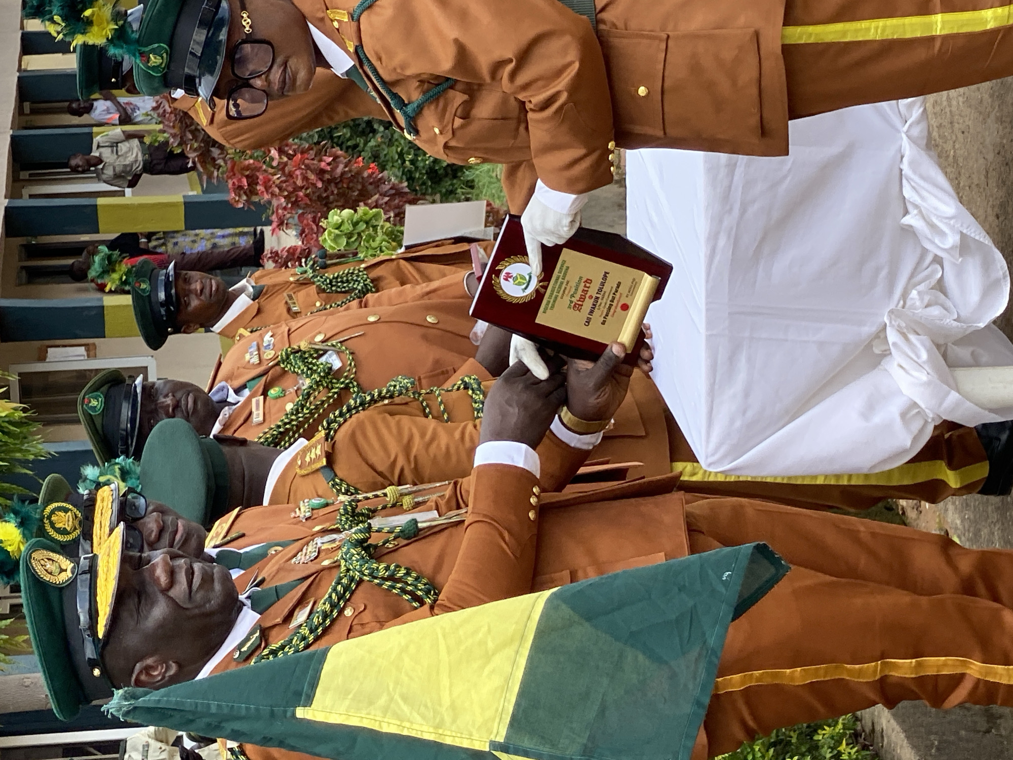 This media file captures a moment at an event in Kaduna, Nigeria. The primary subjects are a group of men in matching brown uniforms with green and yellow accents, wearing green caps adorned with gold emblems. These uniforms suggest a formal or ceremonial occasion, possibly a graduation or award ceremony.
In the foreground, one man is being presented with a wooden plaque by another man also in uniform, who is wearing white gloves. The plaque has text on it, with "Award" clearly visible, along with what appears to be a name and a description. The man receiving the award is looking towards it with a neutral expression. Behind them, other men in similar uniforms are lined up, some appearing to be observing the presentation.
The setting seems to be outdoors, with greenery visible in the background. The presence of a white cloth draped over a surface on the right suggests a formal setup. The lighting indicates it is likely daytime. The overall atmosphere appears formal and celebratory, given the presentation of an award and the uniform attire of the participants. The visible text on the award provides specific details about the event, mentioning "Award," "3rd Position," and a name.