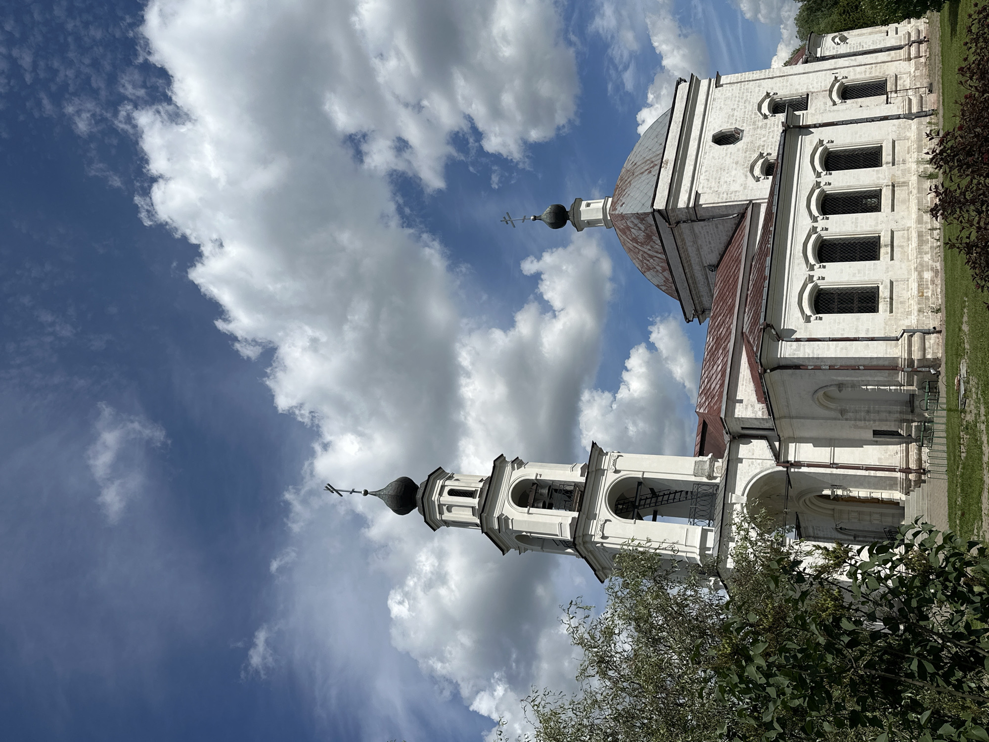 In Erino, Russia, a white church with a prominent bell tower and a domed roof stands against a bright blue sky filled with fluffy white clouds. The bell tower, a multi-tiered structure topped with a dark dome and a cross, features arched openings where bells hang. Adjacent to the bell tower is the main church building, also white, with arched windows and a reddish-brown roof. Lush green foliage from trees and bushes partially frames the left and bottom portions of the image. The scene appears tranquil and idyllic, with no people visible.