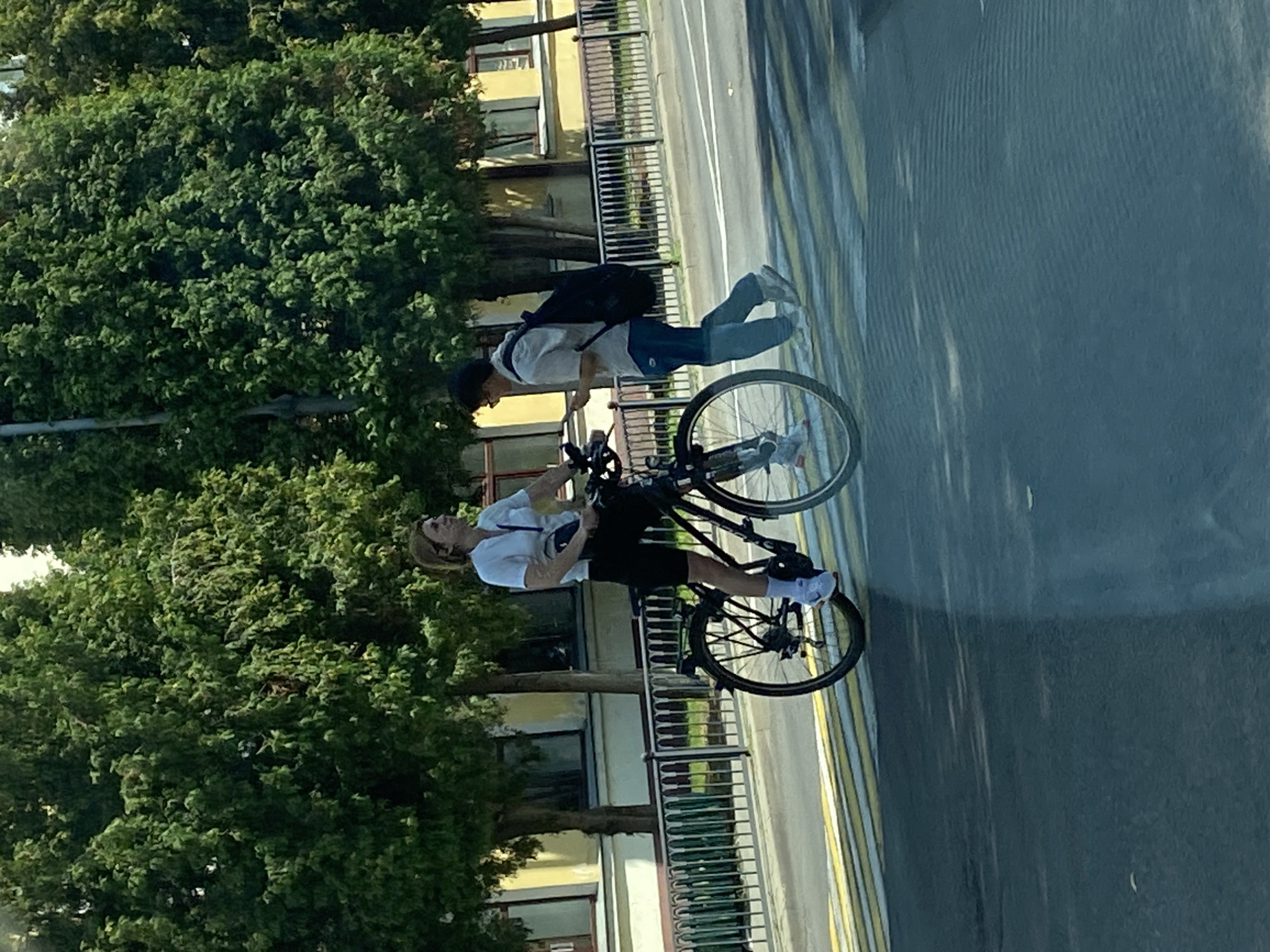 In Brest, Belarus, on a sunny day, a woman on a bicycle is seen at a pedestrian crossing, while another person walks across the street. The woman, with blonde hair and wearing a white short-sleeved shirt and black shorts, is on a black bicycle. She has a black bag around her waist. The other person, wearing a light-colored collared shirt, dark pants, and a backpack, is walking. The background shows trees and a building with yellow walls and dark windows. The pedestrian crossing has yellow and white stripes.