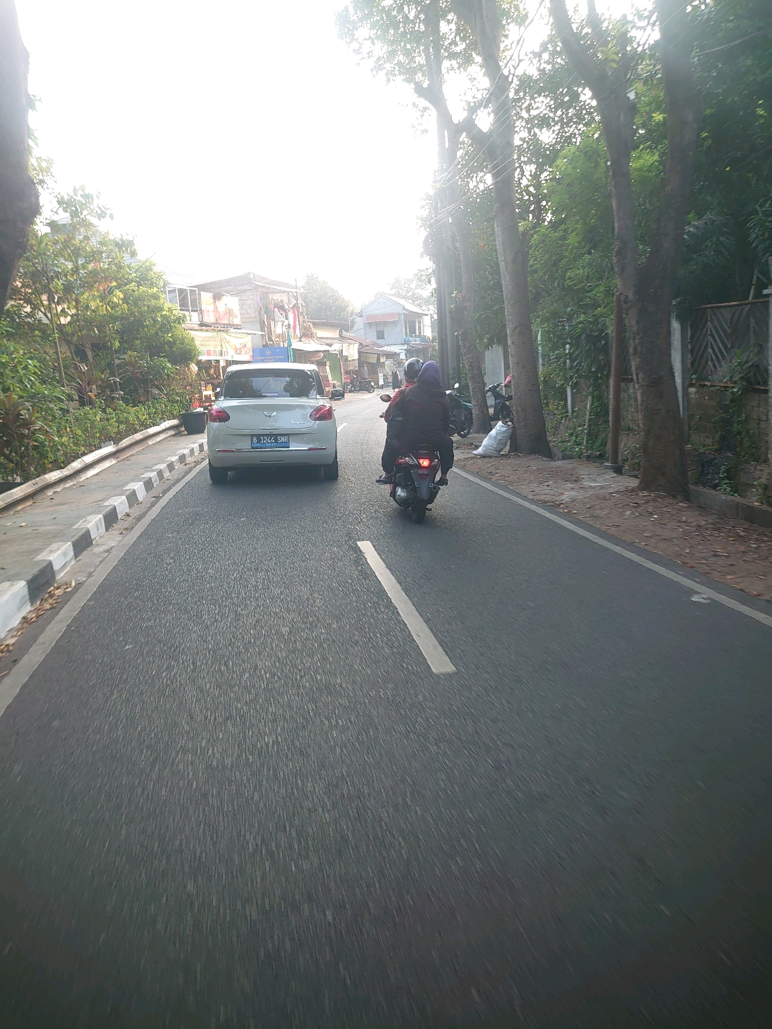 A white car with license plate B 1244 SNR is parked on the right side of a two-lane road. The road is paved with asphalt. There are trees and buildings on the left side of the road. On the right side of the road, there is a sidewalk with a fence. A person is riding a motorcycle behind the car. The sky is bright and sunny. The location is South Jakarta, Indonesia.