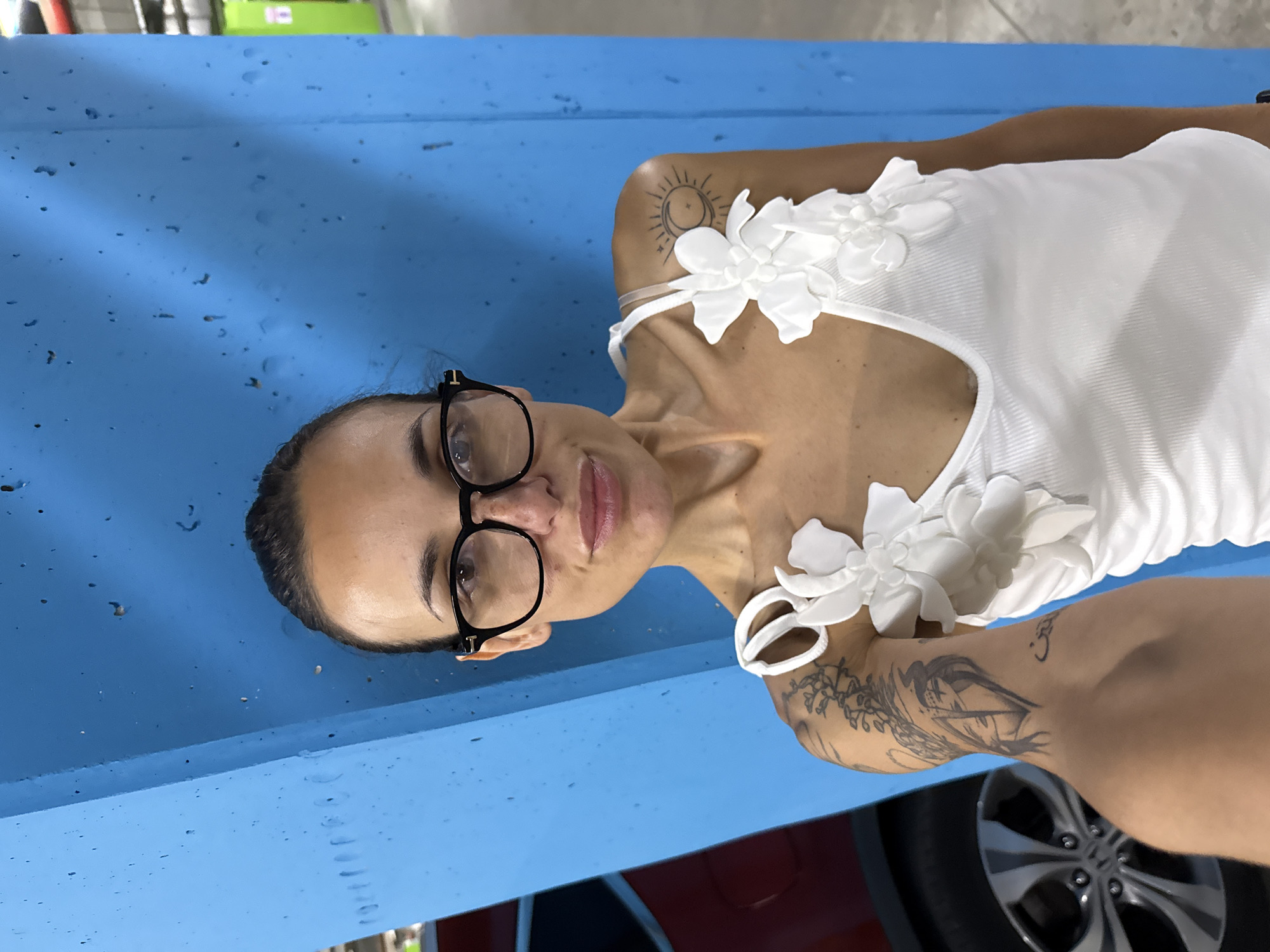 A woman with dark hair pulled back and wearing glasses poses for a selfie in front of a blue concrete pillar. She is wearing a white tank top adorned with white floral appliques. Tattoos are visible on her left arm and shoulder, including a sun and moon design on her upper arm. A portion of a red car and its wheel are visible to the left of the woman. The background appears to be an indoor parking garage with concrete walls and some yellow and green structures in the distance. The specific location of Pleven, Bulgaria, is not evident from the image.
