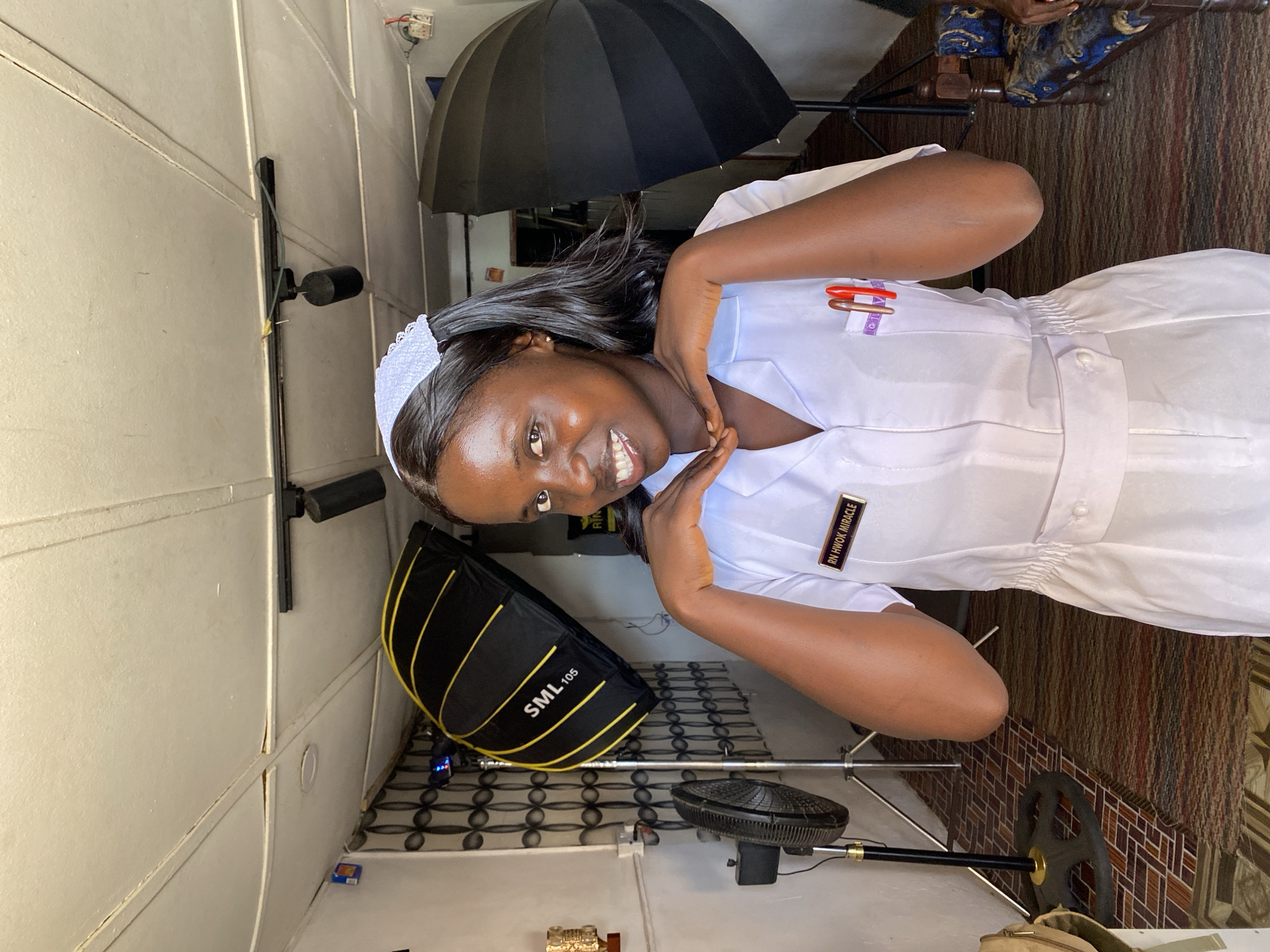 A young woman in a white nurse's uniform with a nurse's cap is smiling at the camera. The name tag on her uniform reads "RN HWOK MIRACLE". She is holding her hands near her face. A large black umbrella light modifier is behind her on a stand, along with another light modifier on the floor. The setting appears to be a simple studio space. The location is Vom, Nigeria.