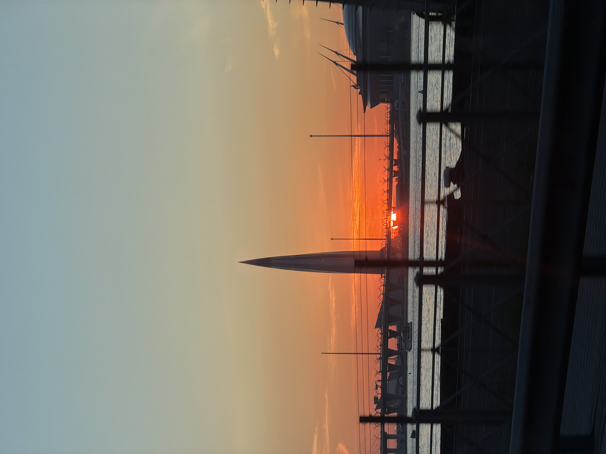 The image shows a sunset in Saint Petersburg, Russia. The scene includes the Lakhta Center skyscraper and the Gazprom Arena stadium, as well as waterfront structures, street lighting, and security fencing. The view is taken from an elevated perspective looking towards the setting sun. The image appears to be taken through or from a building with some structural elements visible in the foreground.