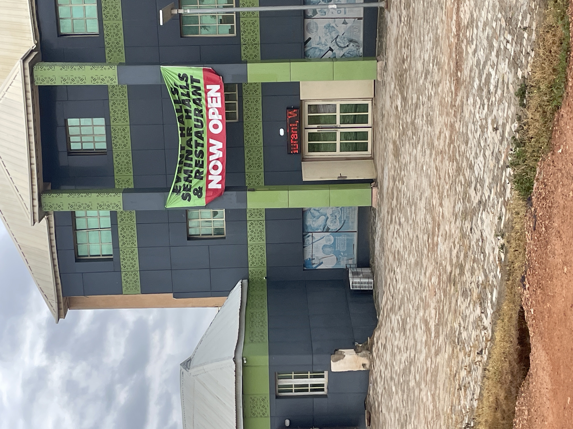 A building in Minna, Nigeria, has a green banner hanging across its facade that reads "EVENT HALLS, SEMINAR HALLS & RESTAURANT NOW OPEN" in large, bold letters. The building appears to be modern with dark gray panels and green accents. There are several windows with a greenish tint. Below the banner, a red digital sign displays "ourani, W". Double doors are open, revealing a dimly lit interior. The ground in front of the building is paved with light-colored bricks. To the left, a smaller, light-colored building with a silver roof is attached to the main structure. An outdoor air conditioning unit is visible near the base of the main building. The sky is overcast with clouds.