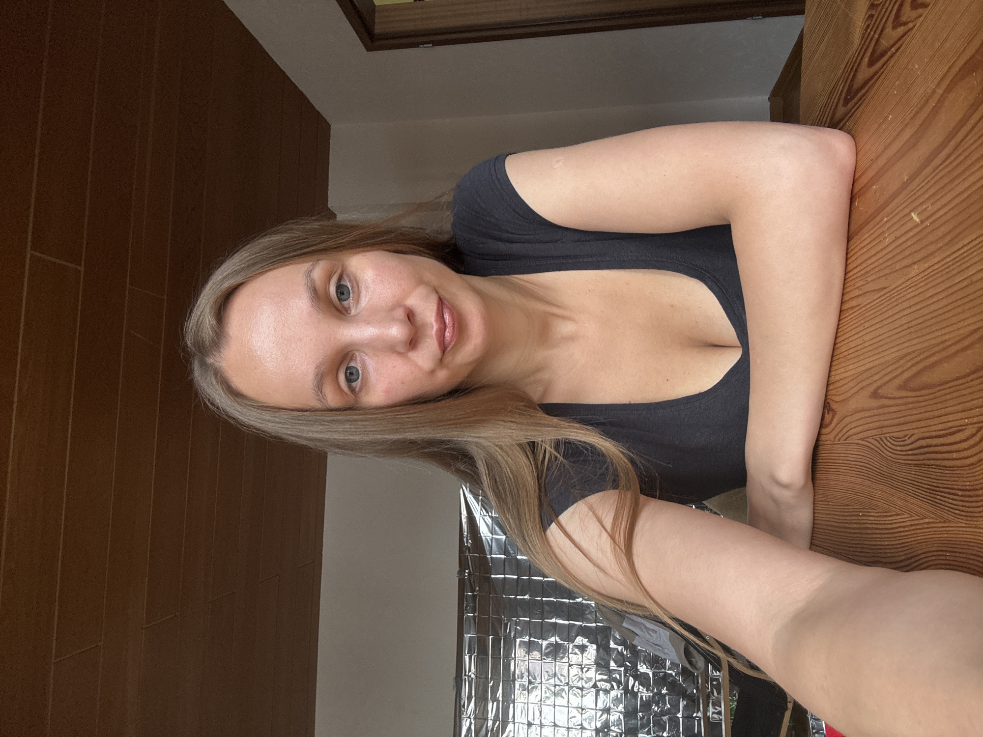 A woman with long blonde hair and blue eyes is sitting at a wooden table, looking directly at the camera. She is wearing a black V-neck t-shirt. Her arms are resting on the table. Behind her, a wall is covered with a shiny, textured material that reflects light, and a wooden ceiling is visible. A doorframe is visible to the right. This scene is not located in Überherrn, Germany.