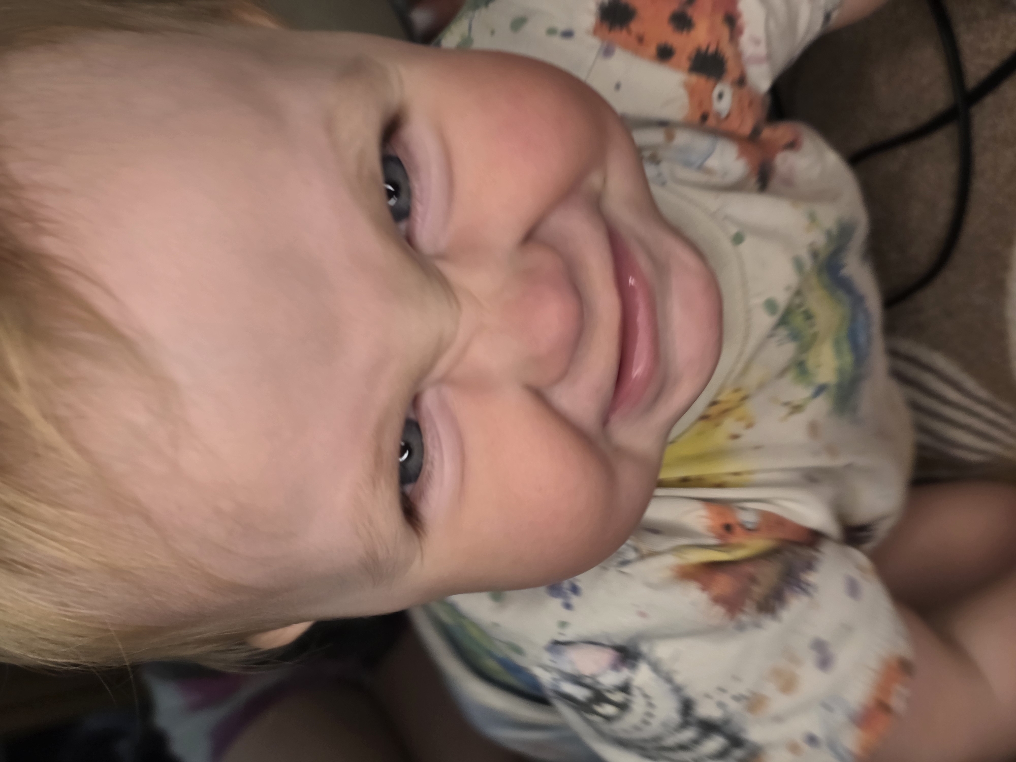 A baby with blond hair, blue eyes, and a fair complexion is looking at the camera, smiling. The baby is wearing a light-colored shirt with a colorful pattern. The background is blurred and appears to be a beige or light brown surface. The location is unknown.