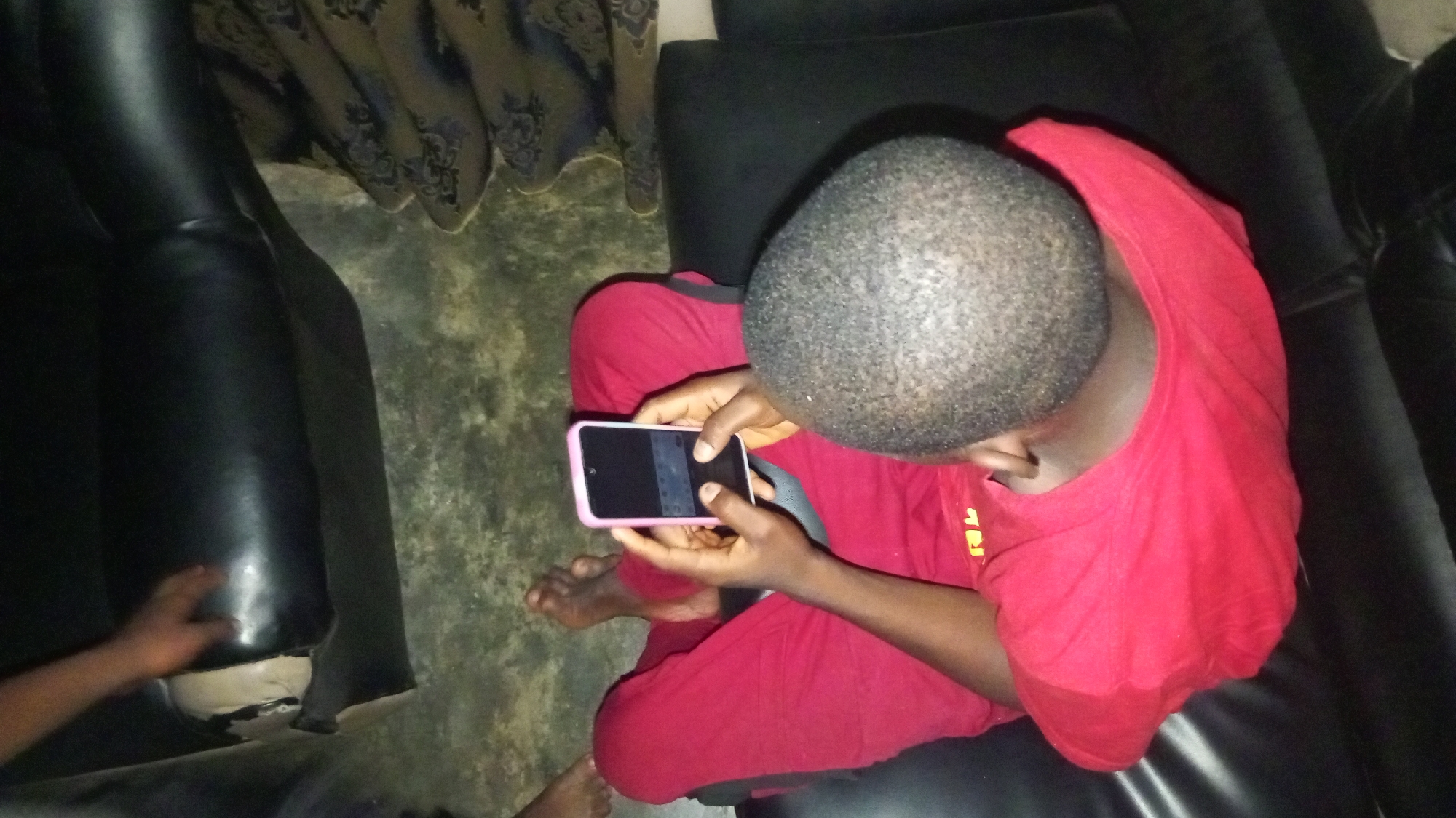 A person in red clothing is sitting on a black leather sofa, looking down at a pink-cased smartphone. The person has short, dark hair. The environment includes a curtain, a patterned floor, and what appears to be another sofa. An arm is visible on the lower left corner of the frame. Location: Unknown, Unknown.