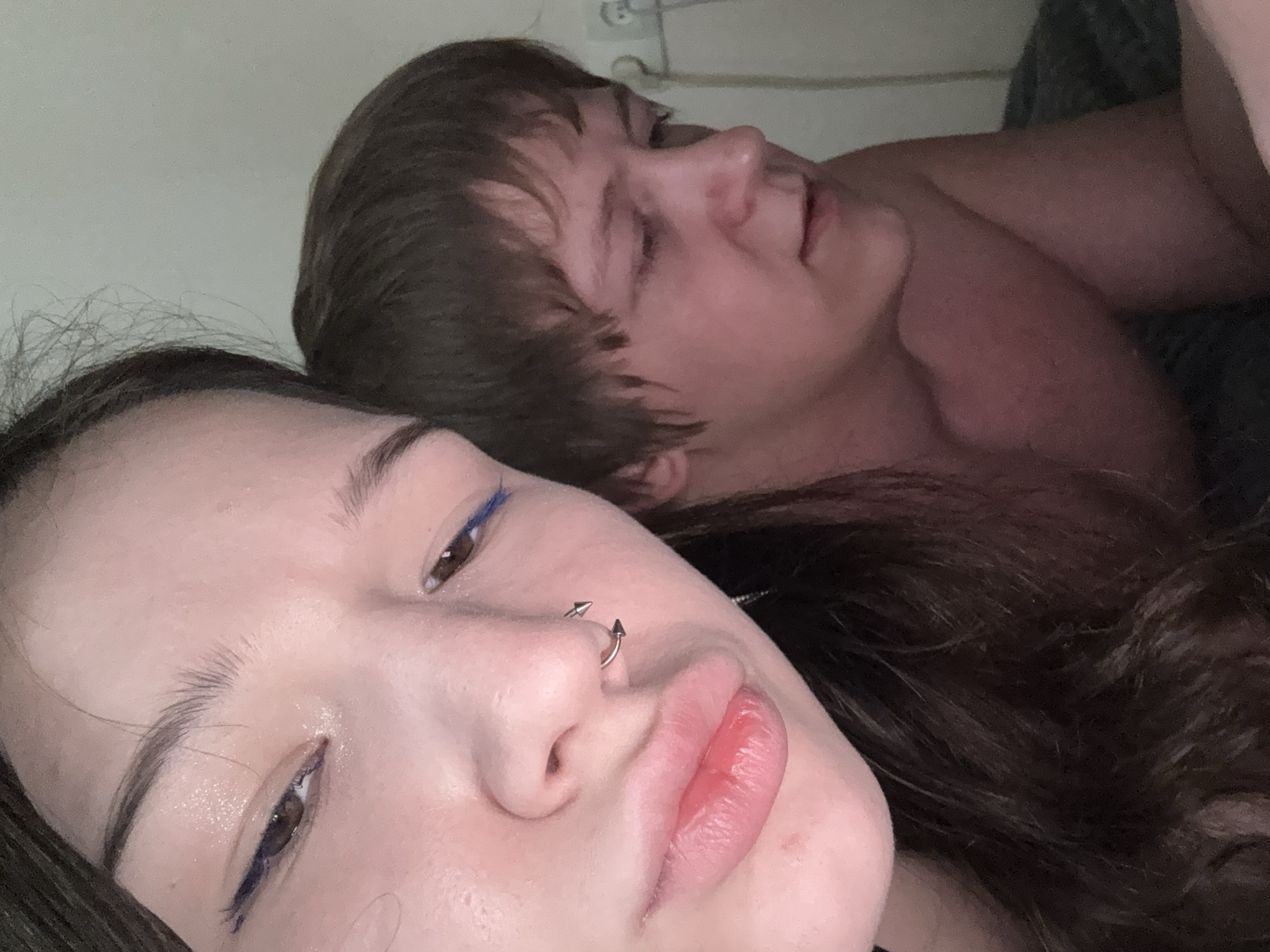 The image is a close-up selfie taken indoors, likely in a bedroom, as indicated by the presence of bedding. Two individuals are partially visible. In the foreground, on the left, a young woman's face is prominently featured. She has dark hair, brown eyes with blue eyeliner, and is wearing a septum piercing. Her expression appears subdued, with her lips slightly parted. To the right and slightly behind her, a young man's face is visible in profile, looking downwards. He has short brown hair and is shirtless. The background is a plain, light-colored wall with what appears to be an electrical outlet visible on the right. There are no discernible landmarks or architectural features that would suggest a specific location within Saint Petersburg, Russia.