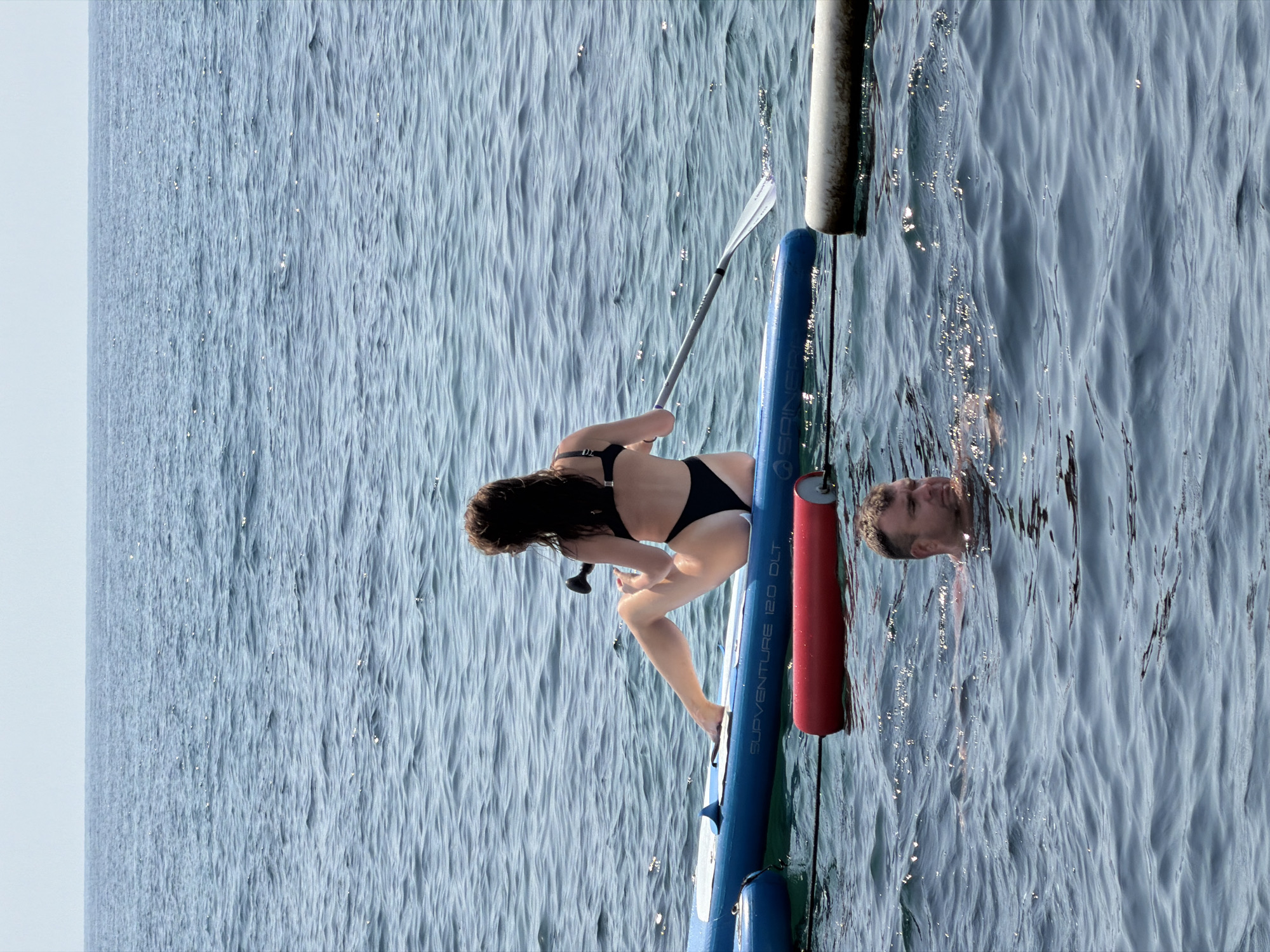 A woman in a black bikini is sitting on a blue paddleboard in the ocean, holding a paddle. She is facing away from the camera, with her legs extended. A man is swimming in the water in front of the paddleboard, with his head and shoulders visible. A red buoy and a white floating object are also in the water. The ocean is calm with small waves. The sky is clear and light blue. The scene appears to be in Budva, Montenegro, a popular tourist destination known for its beaches and coastline.