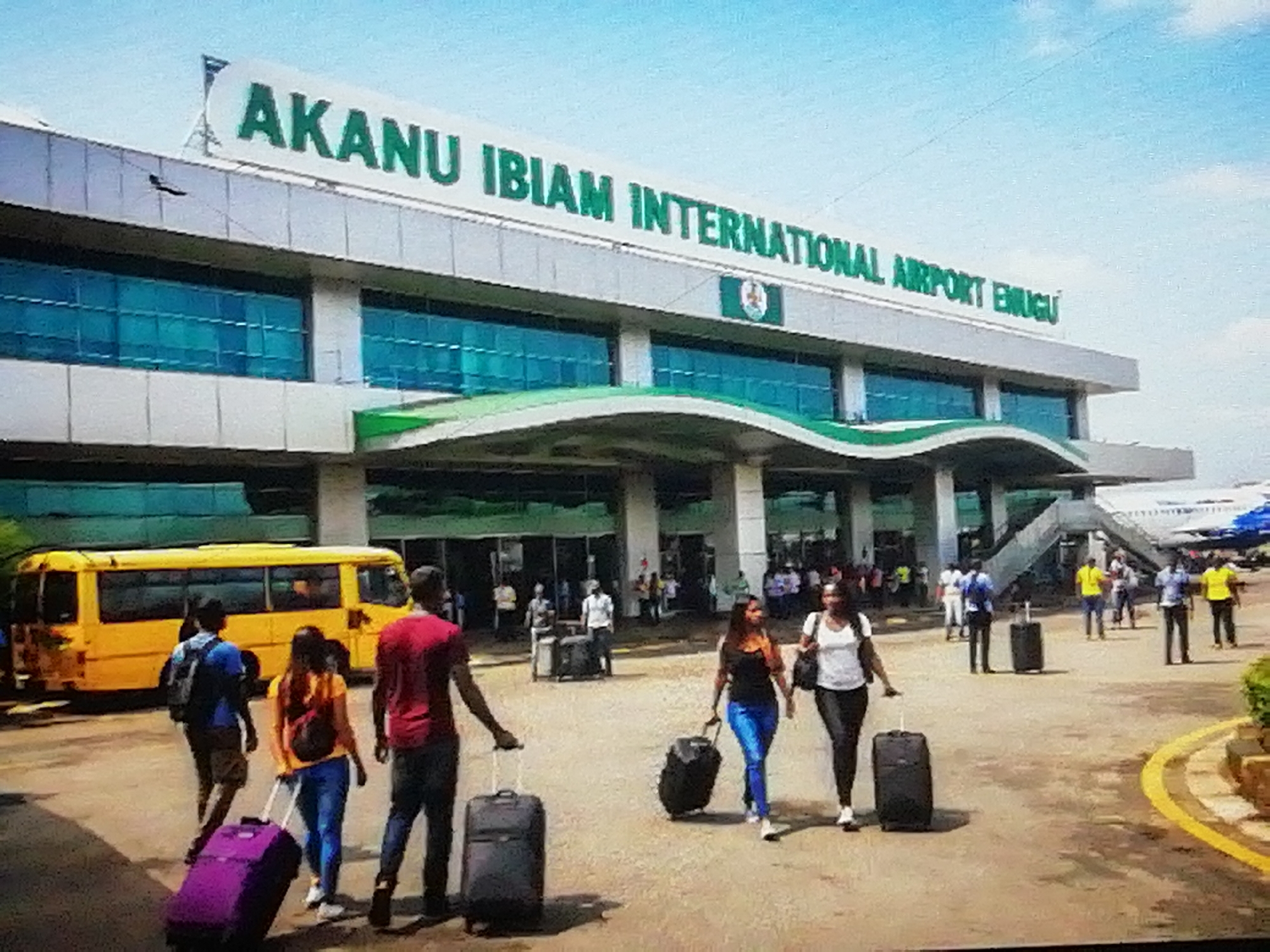 The image displays the bustling exterior of the Akanu Ibiam International Airport in Enugu, Nigeria, under a bright, clear blue sky. The modern airport terminal building is a prominent feature, with its multi-story structure, large blue-tinted windows, and a distinctive green and white arched canopy over the entrance. A large sign affixed to the top of the building clearly reads "AKANU IBIAM INTERNATIONAL AIRPORT ENUGU" in bold green letters. A Nigerian flag is also visible on the building's facade.
In the foreground and midground, numerous people are seen moving around the airport premises. Many individuals are travelers, identifiable by the rolling suitcases they pull, with visible luggage in black, purple, and gray. Some people carry backpacks or shoulder bags. A group of four individuals, including two women and two men, are particularly noticeable in the foreground, walking with their luggage towards the terminal. A bright yellow bus is parked to the left of the entrance, likely serving as an airport shuttle or public transport. In the background, past the main entrance, a section of a large passenger airplane is visible, parked on the tarmac. The paved ground features a distinct yellow curb line. The scene captures the typical activity of an airport, with people arriving and departing on a sunny day.