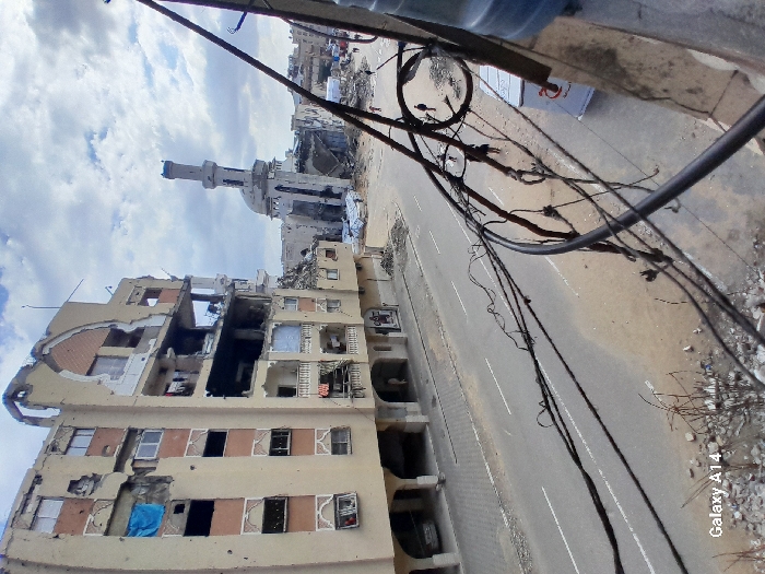 The image captures a wide street scene in Khan Yunis, Palestinian Territory, showing extensive urban destruction under an overcast sky. In the foreground, numerous black electrical wires and cables are strung horizontally across the frame, originating from the right and partially obscuring the view from a higher vantage point, possibly a balcony, where a blue water dispenser bottle is visible on the far right.
To the left, a multi-story building stands significantly damaged, with large sections of its beige facade destroyed, revealing exposed interior rooms, broken windows, and debris. A blue tarp hangs from one of the upper-floor openings. The building's lower level features arched entryways, and a single parked vehicle is partially visible beneath one.
Further down the street, on the right side of the frame, a mosque complex is severely damaged, with its dome partially collapsed and its minaret noticeably compromised. The area around the mosque is littered with rubble and debris. A white banner with red text, likely signage from a relief organization, is visible on the ground near the mosque.
The paved street, with visible white lane markings, runs between the damaged structures and appears relatively clear, though dirt and scattered small debris are present along its edges and on the sidewalks. Several individuals are seen walking on the street and sidewalks in the mid-ground and background, navigating the scene of destruction.