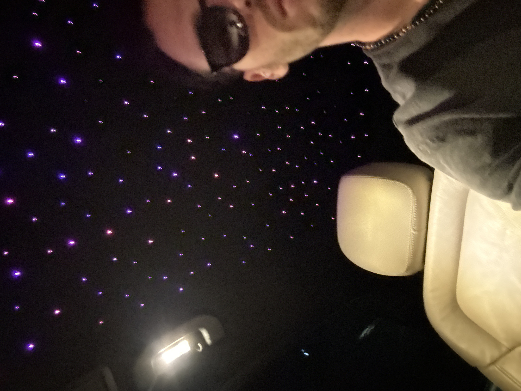 The image shows the interior of a vehicle, possibly a limousine, with a dark ceiling illuminated by numerous small, multicolored lights resembling stars. A person with sunglasses and a beard is visible in the upper part of the frame, wearing a dark-colored shirt. A light-colored leather car seat is also visible. There is nothing in the image to indicate the city of Milan, Italy.