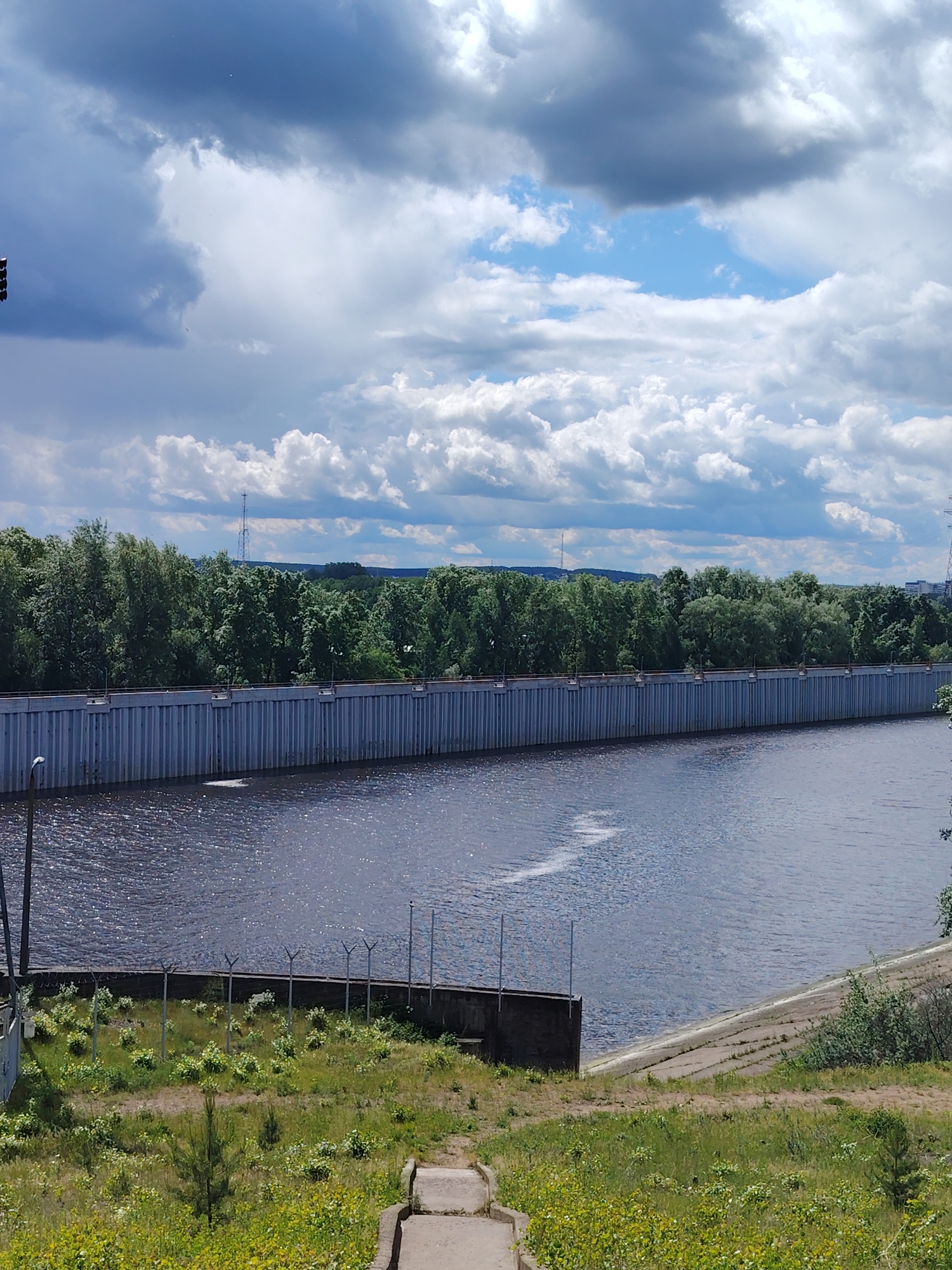The image depicts a water reservoir in the city of Chaikovsky, Russia. The reservoir is enclosed by a long, vertical, corrugated metal wall. Behind the wall, a dense line of trees is visible. Several antenna towers are visible in the background. The sky above is partly cloudy. In the foreground, there is a grassy slope leading down to the water's edge. A short concrete staircase leads down the slope. A low barbed-wire fence runs along the top of the slope.