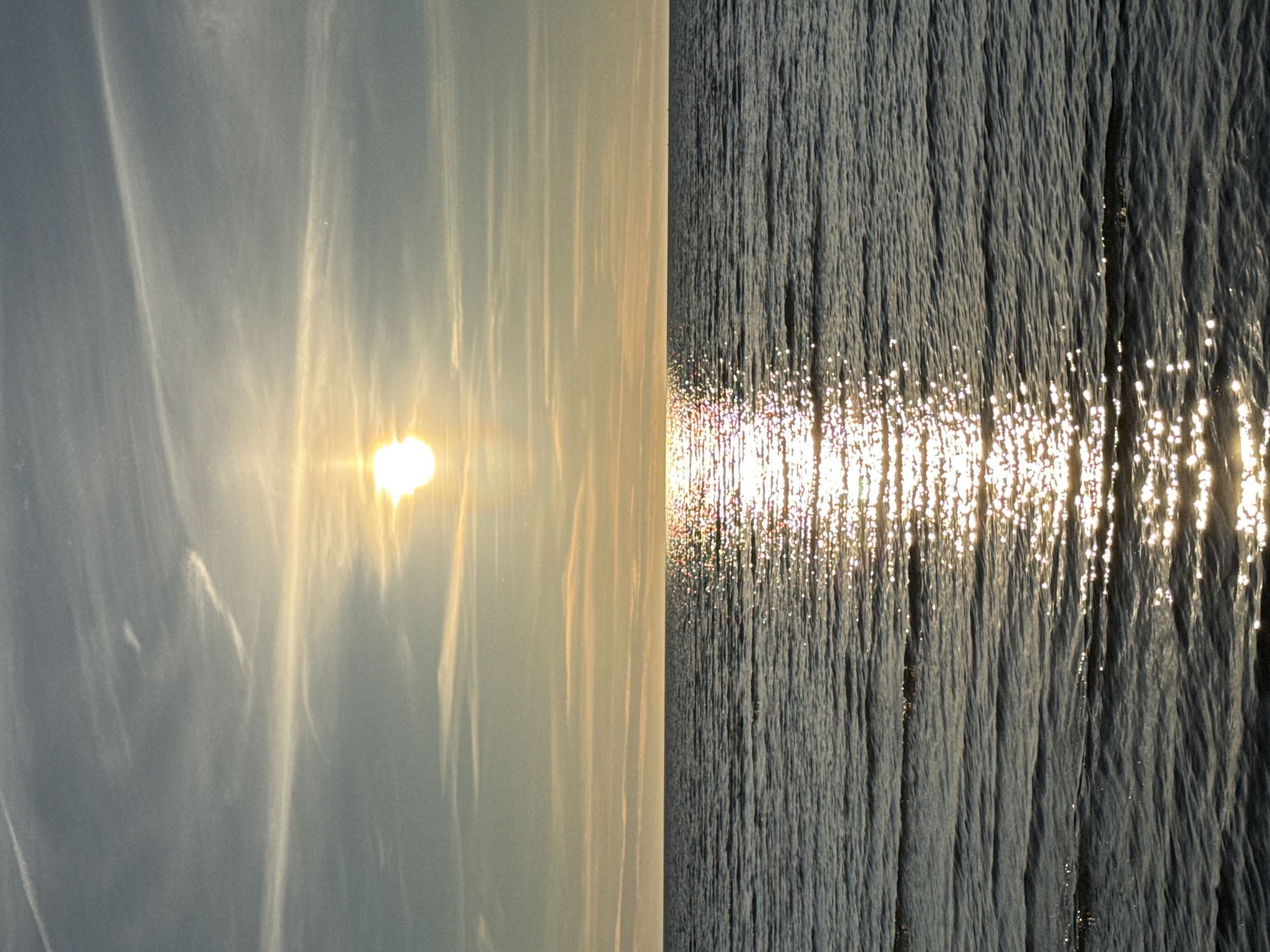 The image depicts a serene seascape at sunset. The sun, a bright orb partially obscured by wispy clouds, casts a shimmering golden reflection across the rippling water. The horizon is clearly defined, separating the pale, cool-toned sky from the darker, undulating surface of the sea. The sky itself is a canvas of subtle color gradients, transitioning from a muted blue at the top to warmer yellow and orange hues near the horizon, with thin, elongated clouds streaking across it. The water exhibits a textured surface due to the gentle waves, with the sunlight creating numerous points of light that dance upon them. There are no people, structures, or discernible landmarks visible in the frame, suggesting an open expanse of water. Given the information provided, it is not possible to determine the specific location within Tallinn, Estonia.