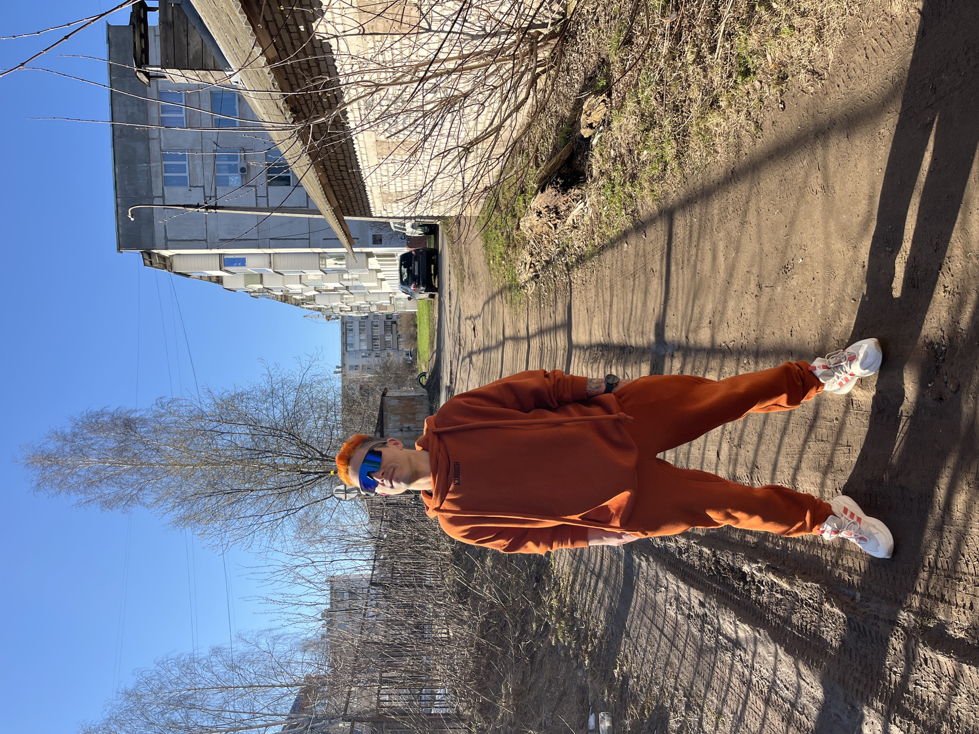 A young man is standing on a dirt path in Chaikovsky, Russia. He is wearing an orange tracksuit, white sneakers with red stripes, and blue mirrored sunglasses. His hair is dyed orange. A building with white brick and a grey roof is behind him. Another building with concrete panels and rows of windows is in the background. Bare tree branches are visible in the foreground. A dark-colored vehicle is parked further down the path. The day is sunny.
