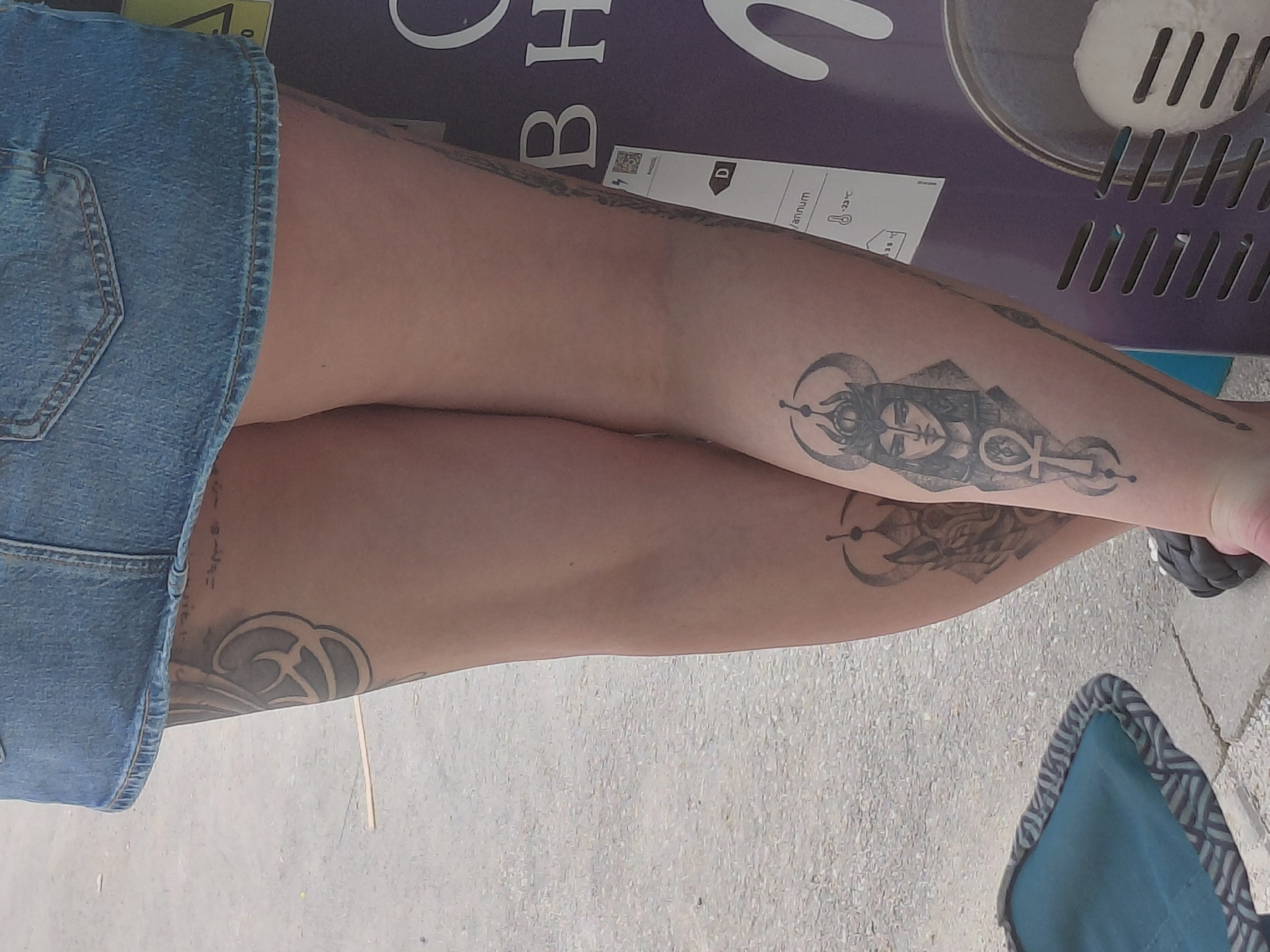 The lower body of a person, wearing a blue denim mini-skirt and dark flip-flops, stands on a textured grey pavement in Unknown, Unknown. The left leg (closer to the viewer) features a tattoo of an Egyptian-style female head wearing a crescent moon crown, with an ankh symbol below it, on the lower calf. A separate, dark, curvilinear tattoo is visible on the upper left thigh. The right leg displays a tattoo of a stylized crowned wolf or creature head with a crescent moon on its lower calf. To the far right, a vertical purple commercial display unit is partially visible, showing white text "O B H" and a white label with a "D" energy rating and "-23°C" indicated. A metal ice cream scoop holding a white spherical object is visible at the bottom of this unit. On the far left, a small, collapsible chair or stool with a blue fabric seat and striped trim rests on the pavement.