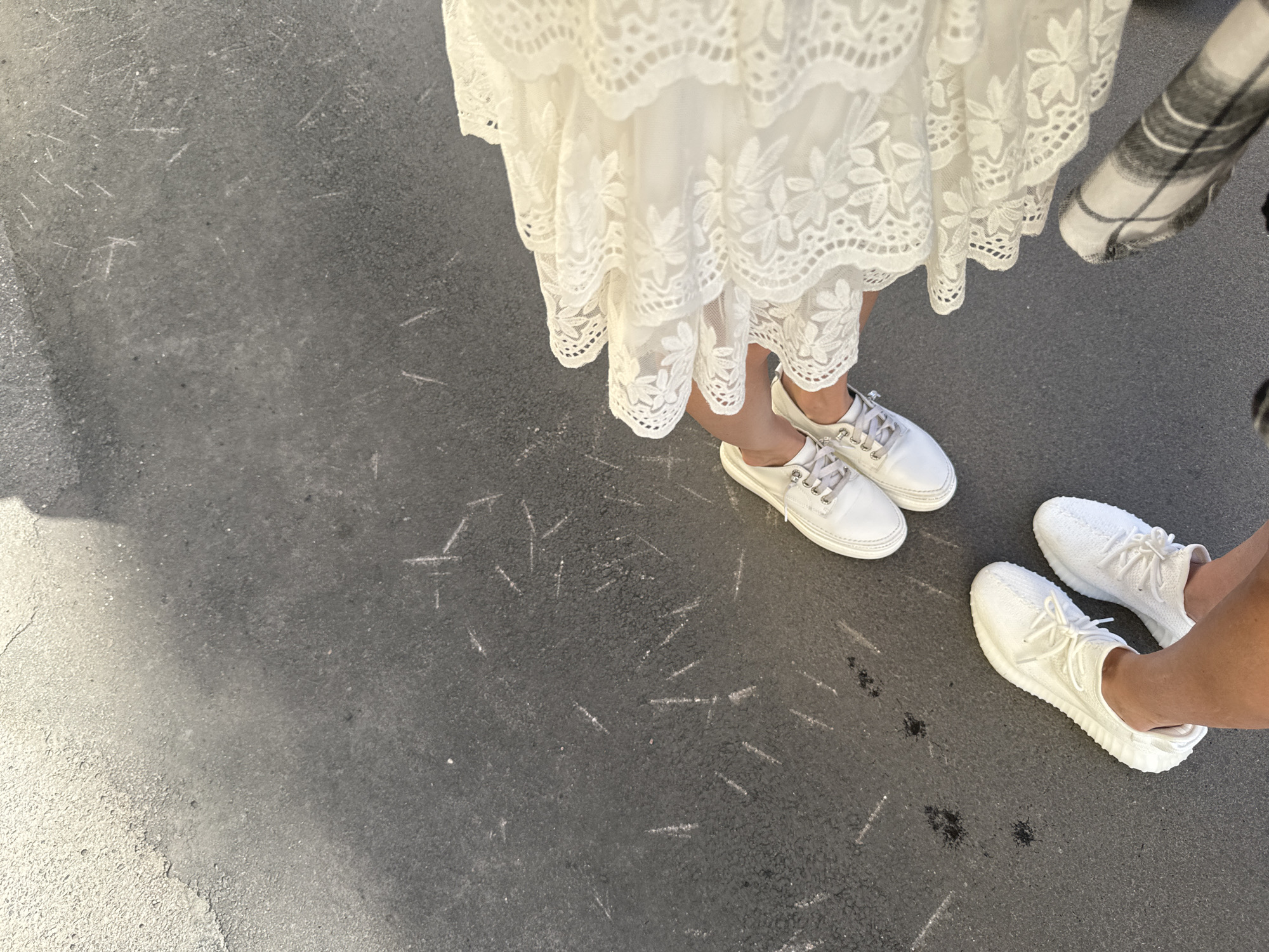 The image shows a top-down view of two pairs of feet on a paved surface. One pair of feet is wearing white sneakers and is positioned towards the bottom left of the frame. The other pair of feet, wearing white sneakers, is positioned towards the top right of the frame and is partially obscured by a white, lacy dress. A person wearing a plaid garment is also partially visible at the bottom right. The ground is dark asphalt with scattered white markings. There is a shadow cast from the top left of the frame. The scene appears to be an outdoor setting in the city of Saint Petersburg, Russia.