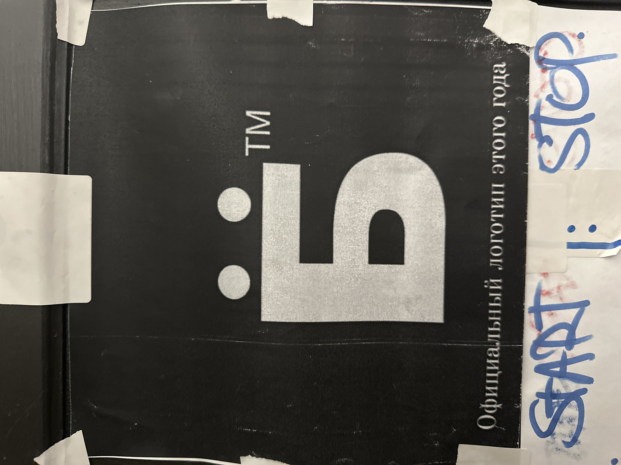 The image shows a black sign with a stylized silver-colored logo resembling a "5" with two dots to the left, and the letters "TM" (trademark symbol) above it. To the right of the logo is text in what appears to be Russian, vertically aligned. The sign is partially covered with pieces of white tape on which the words "START" and "STOP" are written in blue marker. The sign appears to be affixed to a dark-colored surface with a black border. The image has been rotated to show the sign on its side. The city is Новоглаголево, Russia.