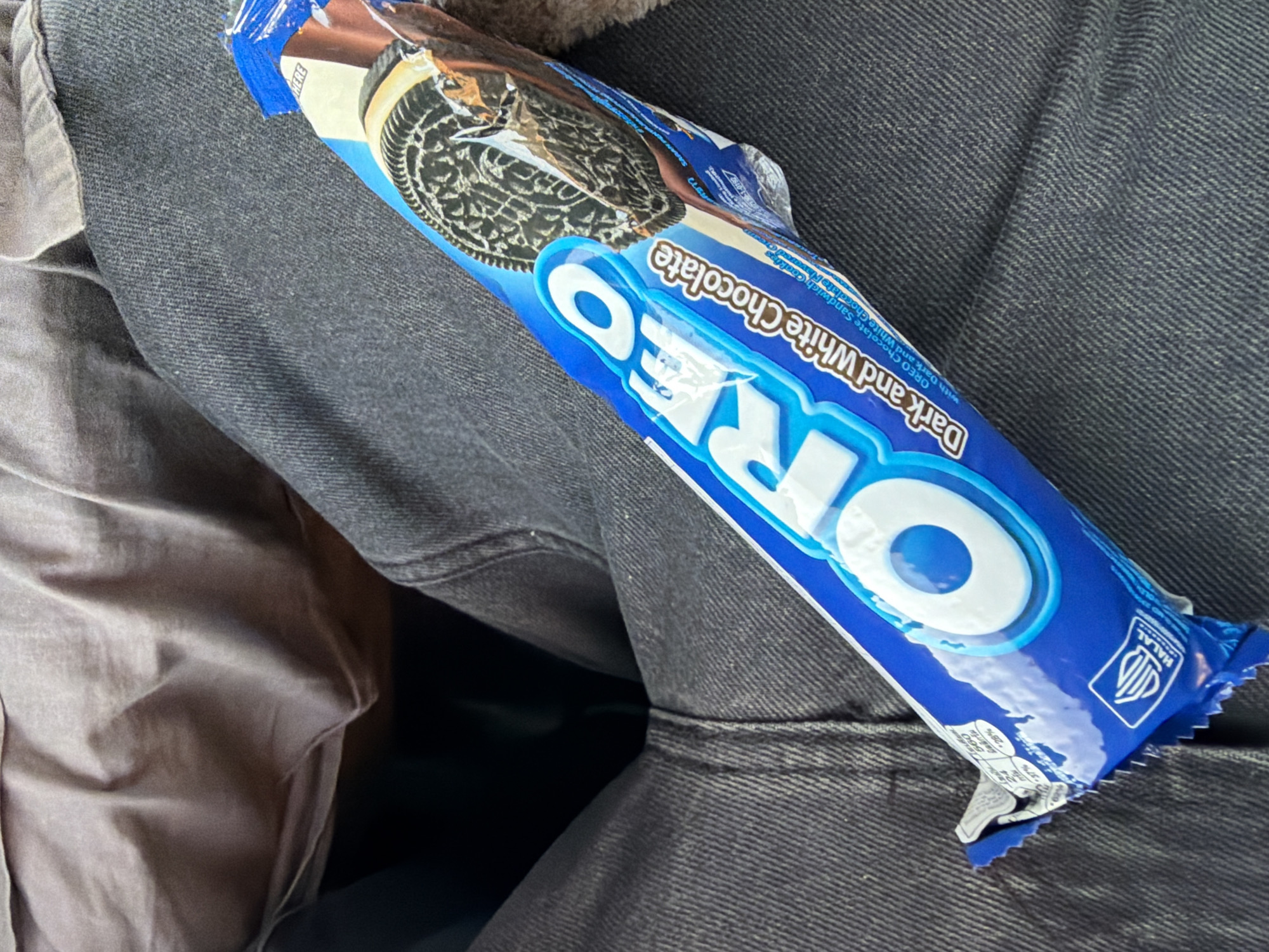 A package of Oreo cookies, specifically "Dark and White Chocolate," rests diagonally across a person's lap, which is clad in dark denim jeans. The person is wearing a dark gray shirt. The background is a blurred expanse of fabric, suggesting the cookies are being held or are on a person's lap while seated. The image is taken indoors. The location is not discernable from the image itself, and therefore cannot be attributed to Gothenburg, Sweden.