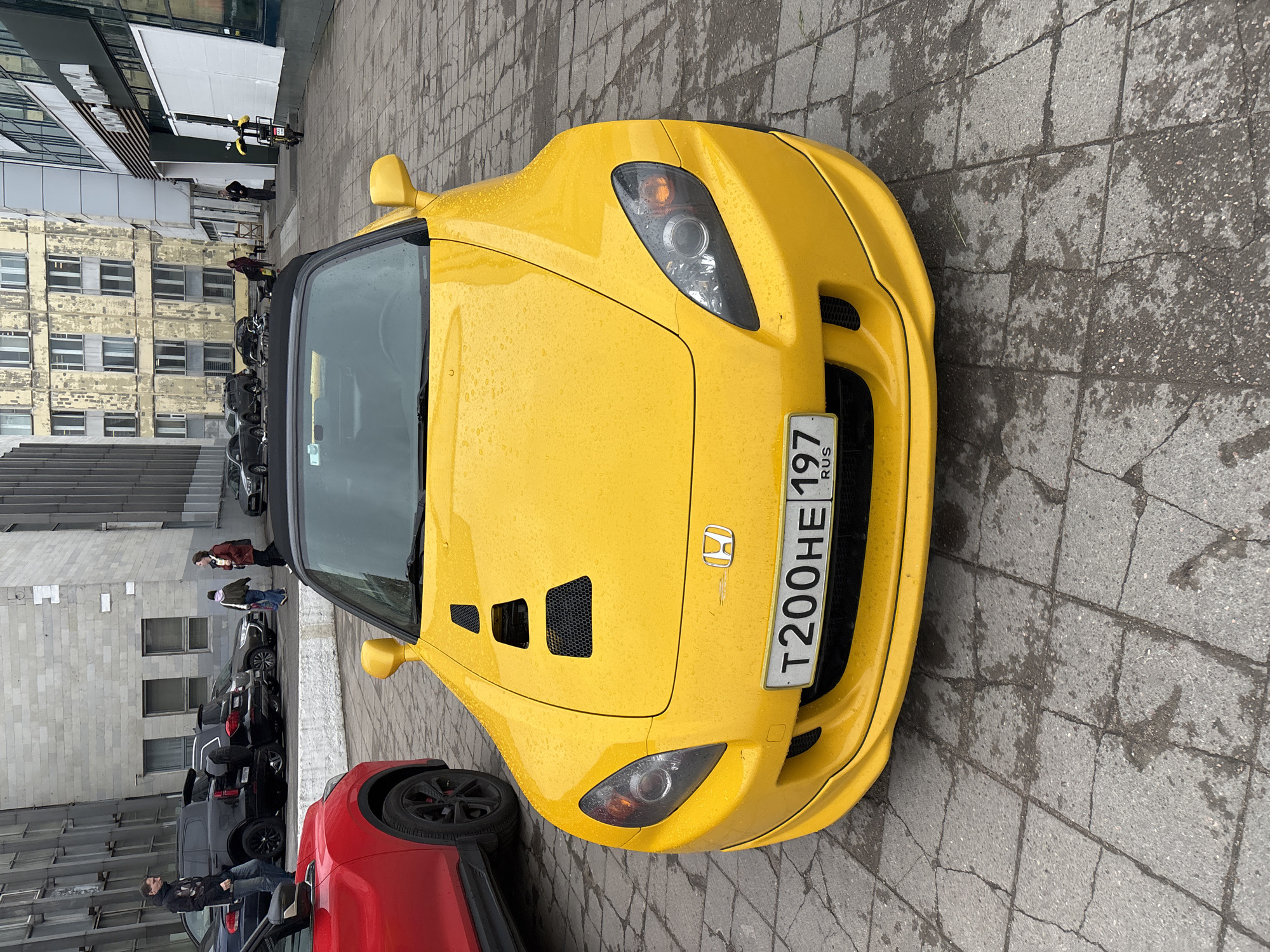 The image shows a yellow Honda S2000 parked on a wet, paved surface near a building. The car has a Russian license plate, T200HE 197 RUS. The location is likely within the city of Saint Petersburg, Russia, based on the license plate code 197. Other cars, including a red one, are parked nearby. Several pedestrians are visible in the background near the buildings. The image is taken from a high-angle perspective.