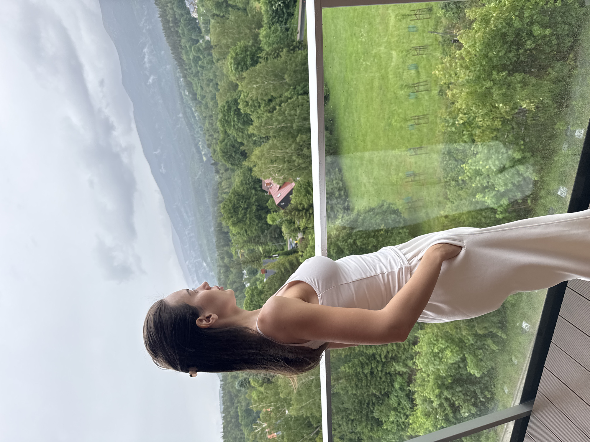 A woman stands on a balcony, captured in profile as she looks out over a scenic, verdant landscape. She is wearing a light-colored, possibly white, sleeveless top and matching long pants, with one hand casually tucked into a pocket. Her dark hair falls over her shoulder, and her gaze is directed slightly upwards and outwards, suggesting contemplation or appreciation of the view.
The setting is an elevated balcony with a modern glass railing and wooden deck flooring. Beyond the balcony, a lush, extensive green area is visible, filled with numerous trees and foliage. A small building with a distinctive red roof is nestled amidst the trees in the middle distance. Further afield, a series of mountains recede into a hazy, overcast sky, indicating either cloudy weather or a misty atmosphere. Directly below the balcony, a large green field or lawn is visible, featuring several uniformly spaced wooden structures that resemble benches or low hurdles.
The image appears to be taken during the daytime, under soft, diffused light from the overcast sky. The overall impression is one of tranquility and natural beauty. There is no visible text in the image.