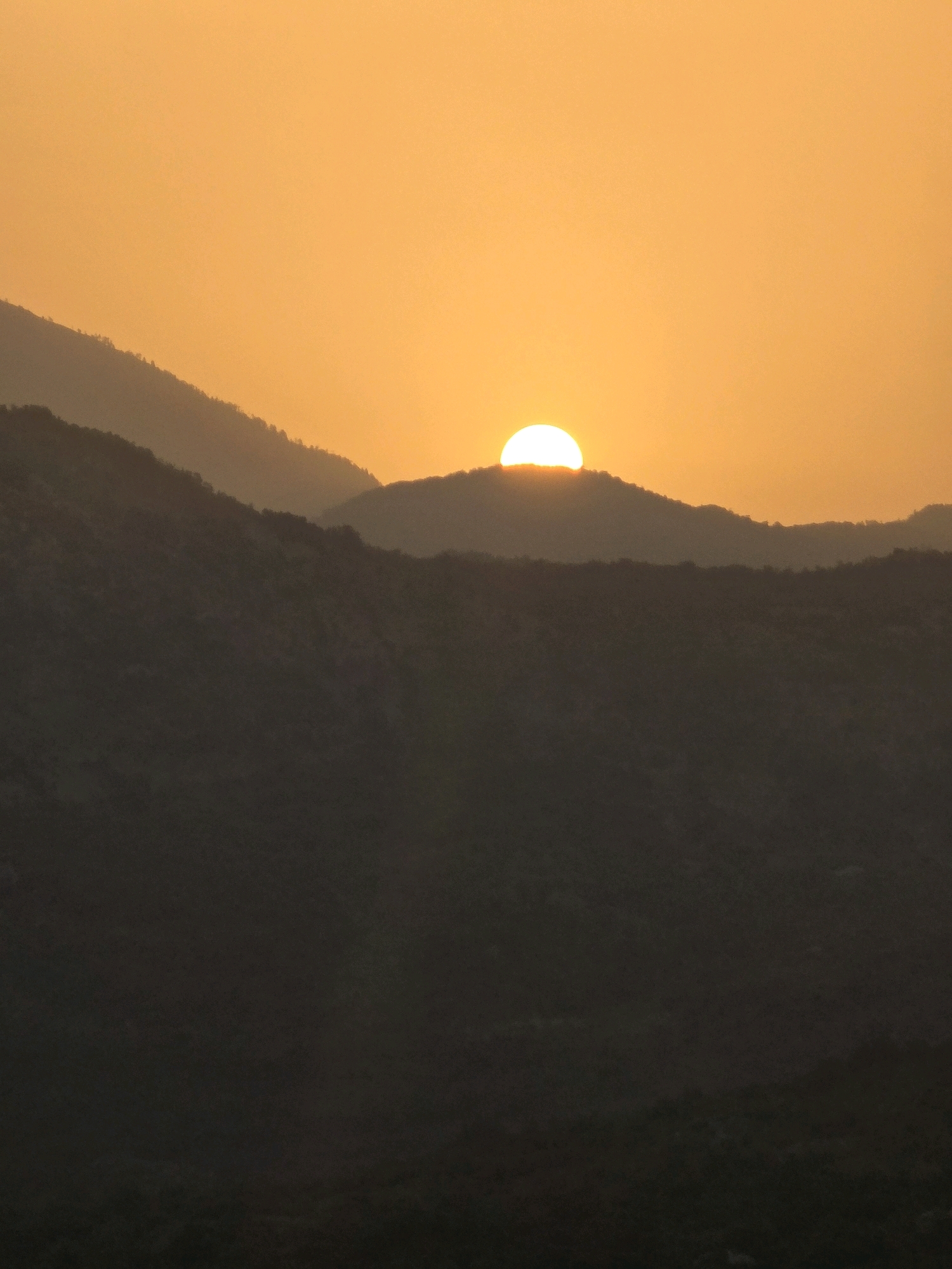 A full frame, vertical, eye-level shot captures a sunset over a mountainous landscape in Medun, Montenegro. The sky is a vibrant gradient of orange and yellow, with the sun, a bright white semi-circle, partially obscured by the crest of a distant mountain range. Silhouetted against the luminous sky are multiple layers of dark, undulating mountains, their peaks softened by the twilight. The foreground consists of darker, more indistinct mountain forms, fading into the shadows at the bottom of the frame. The overall mood is serene and majestic, with the natural beauty of the sunset dominating the scene.