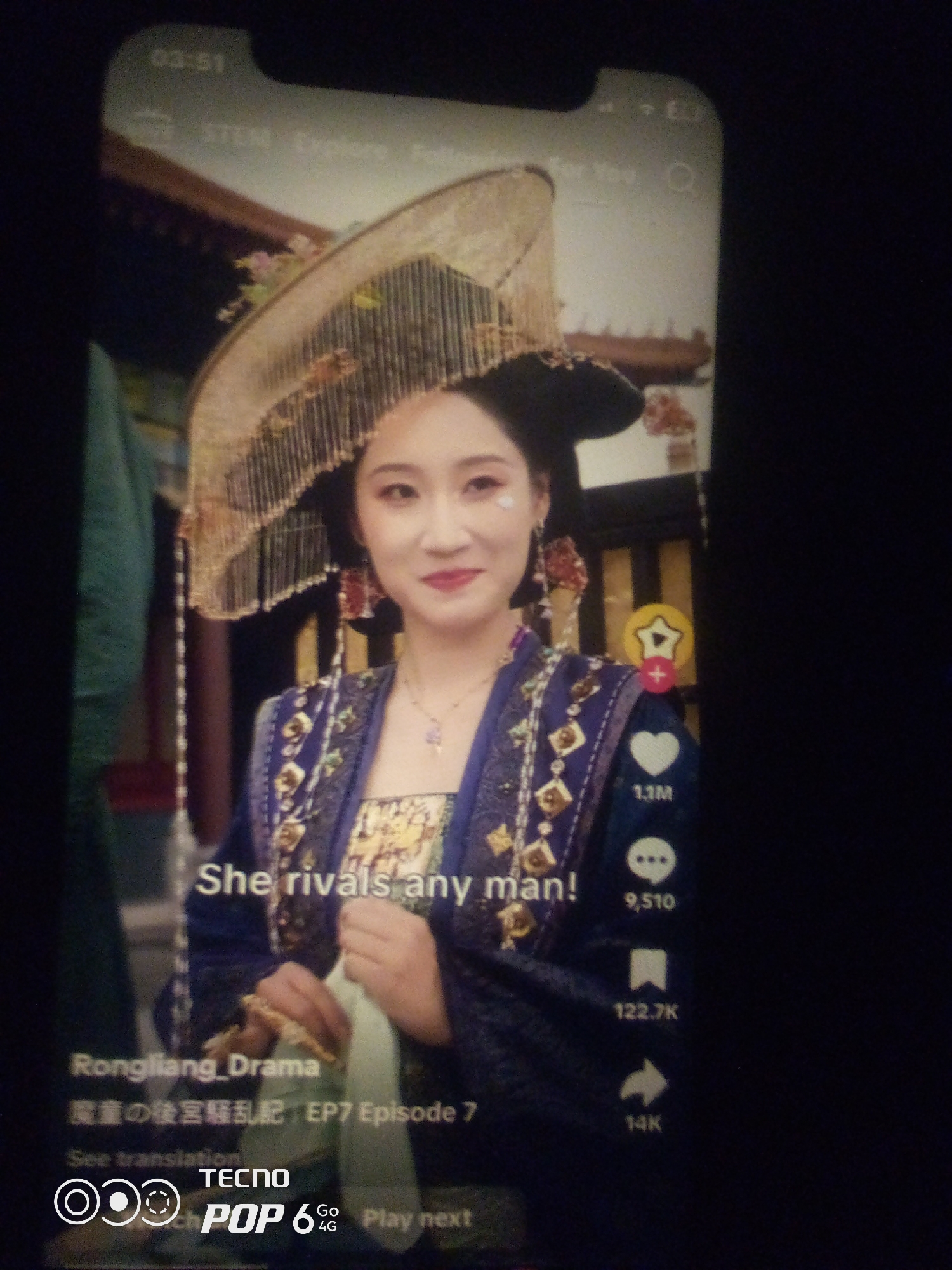 A woman dressed in traditional Chinese attire, including an ornate headdress and a blue embroidered robe, is captured in a medium shot. She is holding a white cloth in her hands and looking towards the camera with a slight smile. The background appears to be indoors, possibly a historical setting, with hints of architectural details. The image is a screenshot from a mobile device, as indicated by the timestamp "03:51" at the top and the presence of interface elements like likes, comments, and share icons on the right side. The text overlay indicates "Rongliang_Drama" and "EP7 Episode 7," suggesting this is a scene from a drama series. There is no visible information to indicate the location as Howayi, Nigeria.