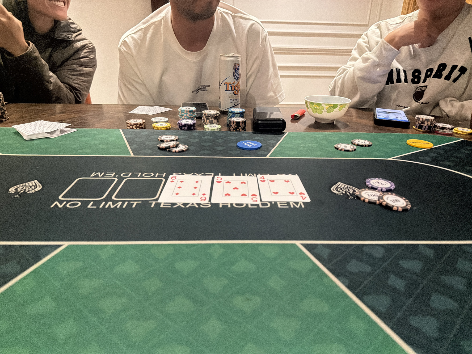 In Xuân Trường - Đà Lạt, Vietnam, a group of individuals are engaged in a game of No Limit Texas Hold'em poker. The scene unfolds around a green felt poker table, where several stacks of poker chips in various denominations are visible. A deck of cards is partially fanned out on the table, and three community cards – the 6 of Diamonds, 5 of Hearts, and 4 of Hearts – are laid face up. One person is wearing a black jacket, another a white t-shirt, and the third a light-colored sweatshirt. A can of Tiger Crystal beer is present, along with a small bowl containing a green and white floral pattern, and a smartphone displaying a screen. The lighting suggests an indoor setting, possibly a home or private room.