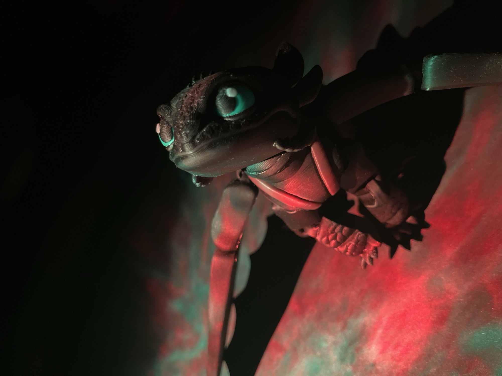 A black, detailed toy dragon figurine with large teal eyes and white pupils is positioned on a surface illuminated with red and green lighting, casting shadows. The figurine is facing to the right, with its head tilted slightly upwards. The surface appears textured and reddish, with streaks of teal light. The background is dark and out of focus. No specific location in Saint Petersburg, Russia, can be identified from the provided image.