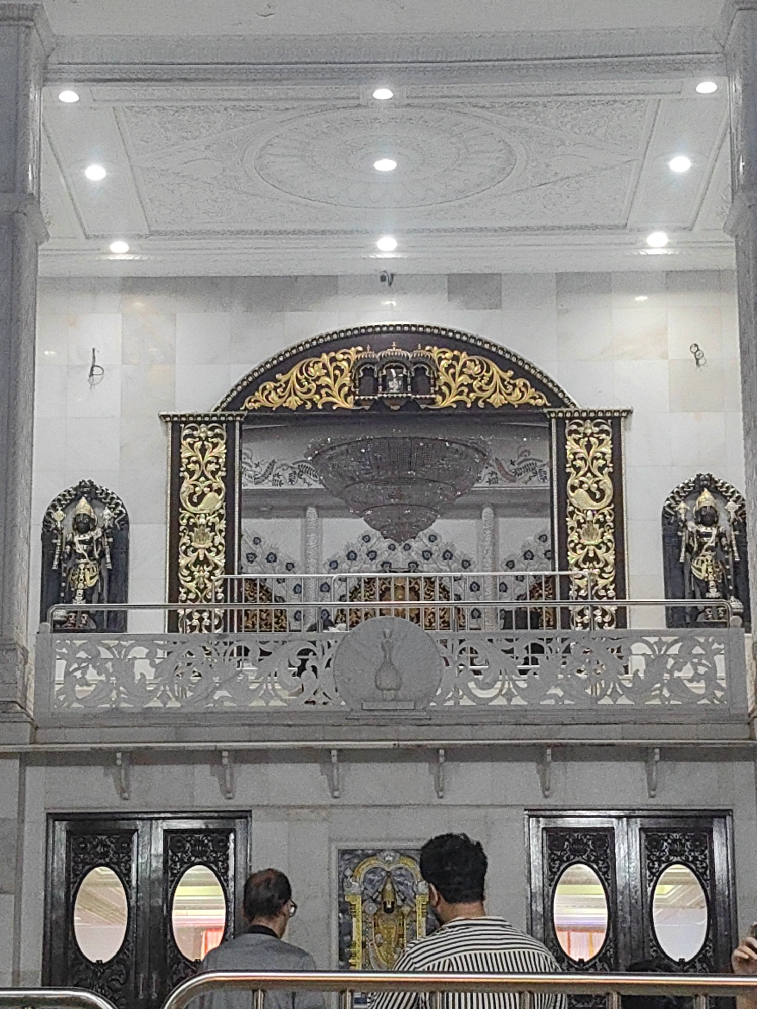 The image shows the interior of a temple, likely in Mira-Bhayander, India, featuring ornate decorations and religious statues. In the foreground, two individuals are partially visible, facing away from the camera. The upper portion of the image reveals a balcony or raised platform with elaborate railings and statues on either side. Behind the balcony, a large, decorative archway frames a chandelier and a patterned wall. The ceiling is adorned with intricate plasterwork and recessed lighting. The lower part of the image shows large, dark wooden doors with oval-shaped windows. The overall scene is one of grandeur and religious significance.