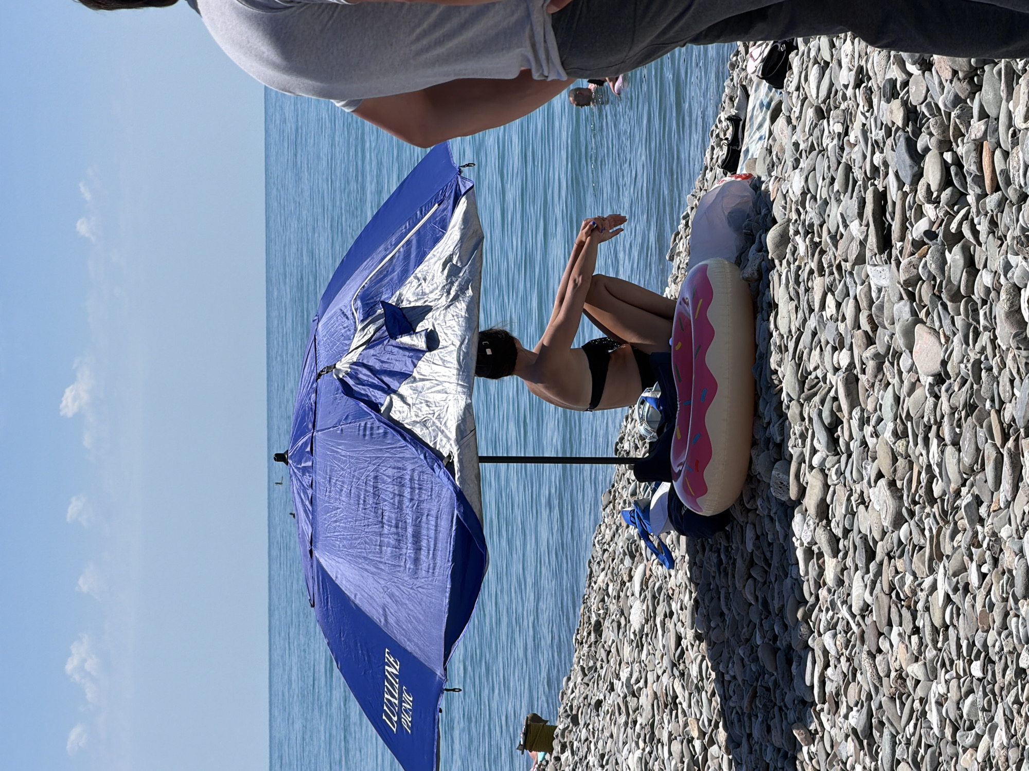 A medium shot captures a beach scene with a woman sitting under a blue and white umbrella on a pebble shore. She is wearing a black bikini and has her knees bent, with her hands resting on her legs. In front of her is a pink inflatable donut. To the right of the frame, a man in a grey t-shirt and grey pants stands with his back to the camera, looking towards the ocean. The ocean is calm and blue, with a few people visible in the water. The sky is clear and bright blue with a few wispy clouds. The ground is covered in smooth, grey pebbles. The scene appears to be in Sirius, Russia, given the context of the prompt.
