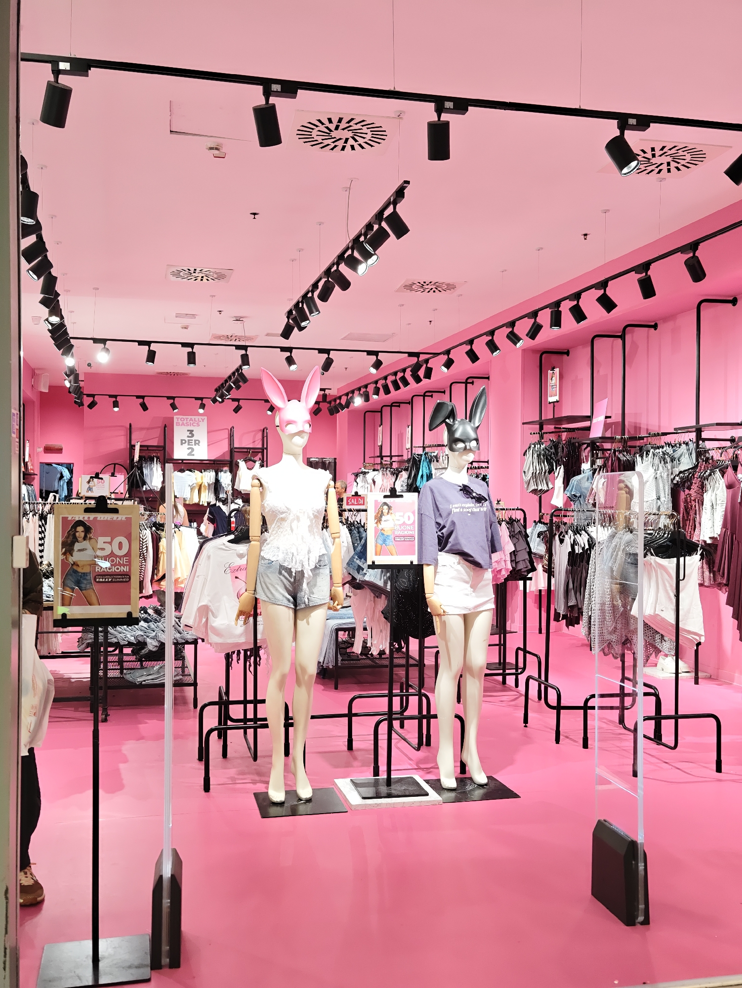 The image depicts the interior of a brightly lit clothing store with predominantly pink walls and flooring. Two mannequins are prominently displayed at the front, both wearing novelty animal-ear masks and fashionable outfits. The mannequin on the left is wearing a white lace top and denim shorts, while the mannequin on the right is in a purple t-shirt and white skirt. Behind the mannequins, clothing racks filled with various garments are visible, illuminated by numerous black track lights mounted on the ceiling. A promotional poster advertising a "50% ONE PRICE" sale is displayed near the left mannequin. The store appears to be located in the city of Nola, Italy, as indicated by text on a sign that reads "NUOVA COLLABORAZIONE" and mentions "NOLA".