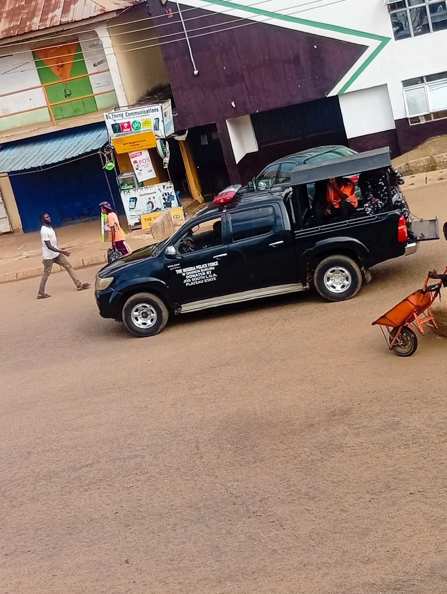 In Bukuru, Nigeria, a black police van with a siren light on top is parked on a paved road. Text on the side of the van reads "THE NIGERIA POLICE FORCE" and further details about its division and donation. A person in an orange vest is visible in the back of the van. Two individuals are walking on the sidewalk in the background, one in a white t-shirt and the other in an orange t-shirt carrying a bag. Adjacent to the road, a store with signs that include "Young Communications" and "POS" is visible. An orange wheelbarrow is positioned on the side of the road. The environment appears to be an urban street with buildings lining one side.