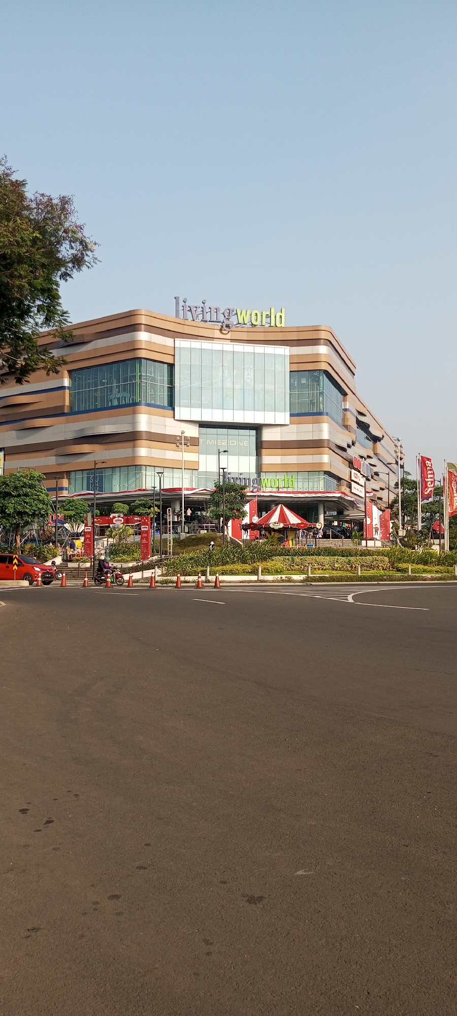 The image shows the Living World mall in Lambangjaya, Indonesia, on a clear day. The modern, multi-story building features a distinctive facade with horizontal bands of gold, white, and blue. Large glass windows are prominent throughout the structure. Above the main entrance, the "living world" logo is displayed in green lettering. In front of the mall, there is a landscaped area with trees and bushes. A red car is parked on the left side of the road, and a person is riding a motorcycle on the right. Several orange traffic cones are placed along the roadside, indicating some form of temporary closure or traffic management. The road in the foreground is asphalt with white lane markings. The sky is a pale blue, suggesting it is daytime.