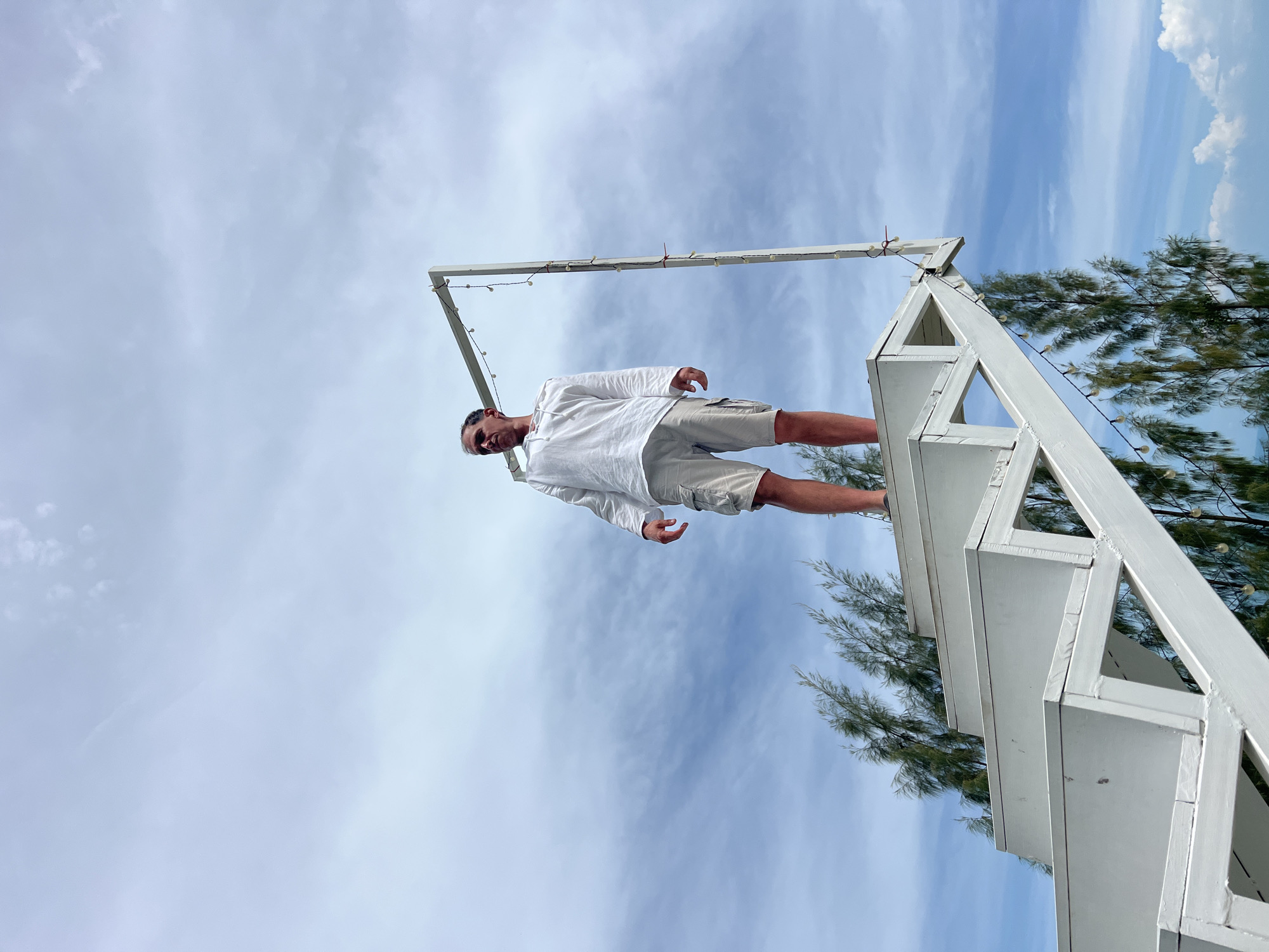 A man stands on a white, multi-level structure, possibly a stage or display. He is wearing a white long-sleeved shirt and light-colored shorts. The structure has a frame adorned with small lights. To the right of the man, there are green trees with thin leaves. The background is a cloudy blue sky. The image is taken from a low angle, looking upwards at the man and the structure. The man is looking towards the camera with a slight smile. The lighting suggests it is daytime, and the weather appears to be clear with some clouds. The location is Baan Thurian, Thailand.