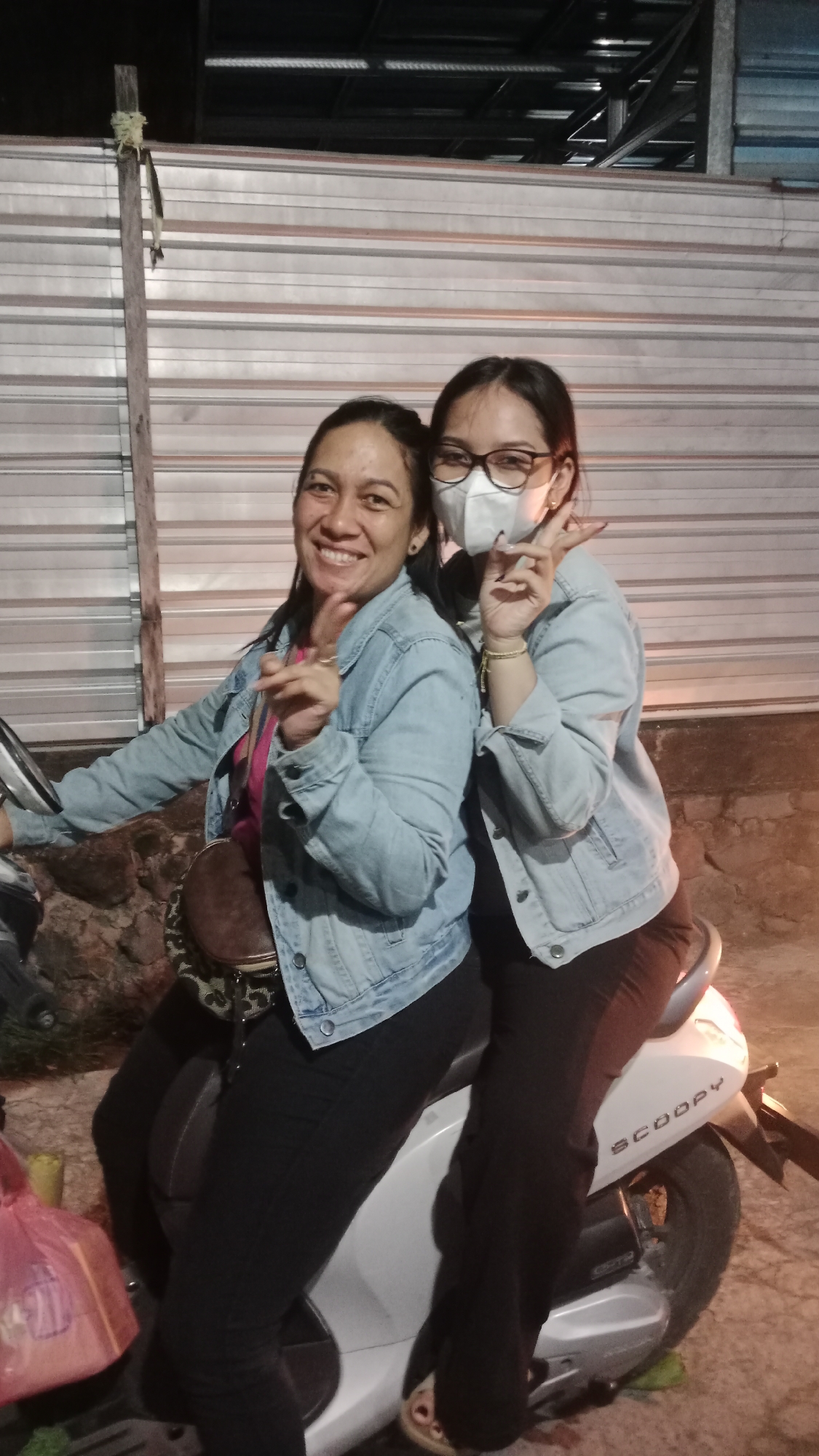 Two women are posing for a photo on a white scooter in front of a corrugated metal wall. The woman in the foreground is wearing a light blue denim jacket, a pink shirt, and black pants. She has dark hair pulled back and is smiling, holding up two fingers in a peace sign. The woman behind her is wearing a similar denim jacket and black pants, a white face mask, and glasses. She is also holding up two fingers in a peace sign. The scooter has "SCOOPY" written on its side. A pink plastic bag is visible on the left side of the image. The lighting suggests it is evening or nighttime, with artificial light illuminating the scene. The background wall has horizontal lines and appears to be part of an outdoor structure. The setting is likely a residential area or an informal commercial space in Denpasar, Indonesia.