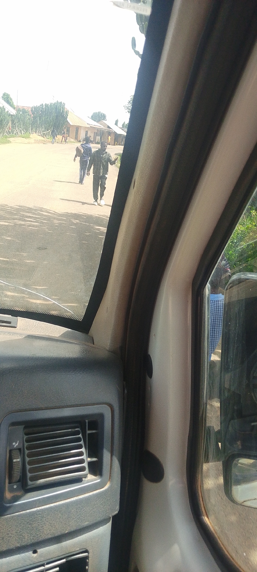 A group of people walk along a paved road in what appears to be a rural or semi-urban setting. The scene is captured from inside a vehicle, with a portion of the dashboard and windshield visible in the foreground. Tall, slender plants, resembling cypress trees, line the left side of the road, interspersed with some buildings that have corrugated metal roofs.
Two men are walking towards the camera. The man on the right is wearing a checkered shirt and dark pants, and appears to be talking with his hands outstretched. The man on the left is walking away from the camera, carrying a bag over his shoulder and wearing a dark hooded sweatshirt and striped pants. Further down the road, other figures are visible, including a person in a green uniform walking away from the camera.
The day appears to be bright and sunny, with clear skies and shadows cast on the road, suggesting it is likely midday. The presence of the buildings, the style of dress of the individuals, and the overall environment point towards a location in Nigeria, specifically Bukuru. There are no visible text elements in the image. The overall impression is one of everyday activity in a community.