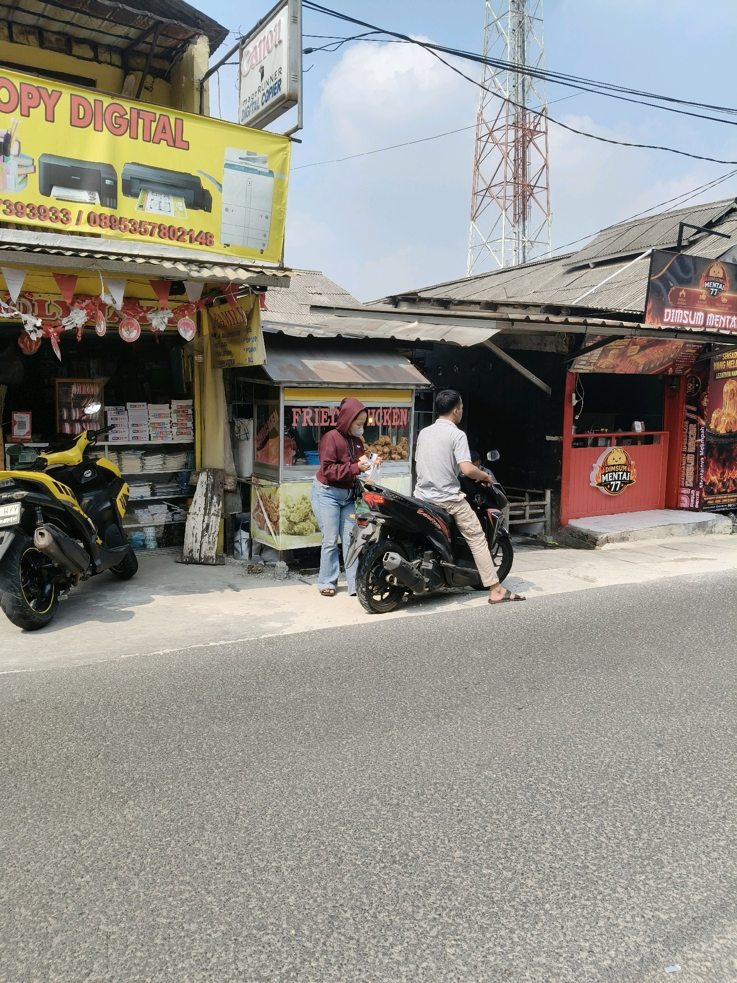 In South Tangerang, Indonesia, a street scene unfolds. A yellow and black motorcycle is parked on the left. Next to it is a storefront with a yellow banner that reads "COPY DIGITAL" and displays images of printers. Above this banner, a white sign with "Canon" and "imageRUNNER DIGITAL COPIER" is visible.
To the right of the copy shop, a food stall is set up, advertising "FRIED CHICKEN" with pictures of fried chicken. A person wearing a maroon hoodie and blue jeans stands near the food stall, facing away from the camera, holding what appears to be a paper bag.
Further down the street, another food stall with red accents displays a sign with "DIMSUM MENTAI 77". A black motorcycle is parked in front of this stall, with a person wearing a grey t-shirt and light-colored pants sitting on it, facing left. This person is looking towards the food stall.
The road in the foreground is asphalt. In the background, a tall communication tower is visible against a bright sky with scattered clouds. The buildings have corrugated metal roofs.