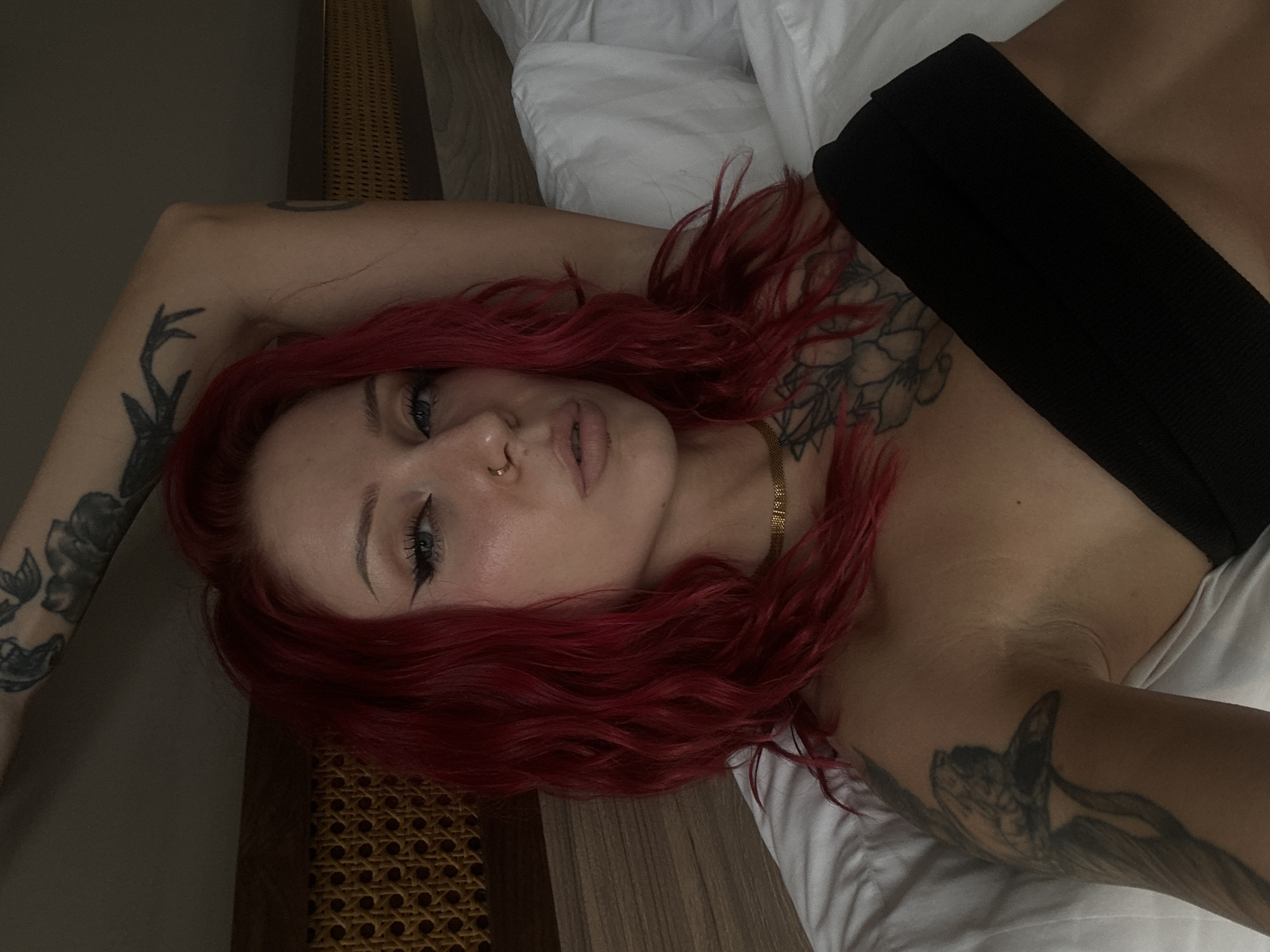 A woman with red hair is lying on a bed in a room with wood paneling. She has a black top on, a gold necklace, and tattoos on her arms and chest. The location is Unknown, Indonesia.