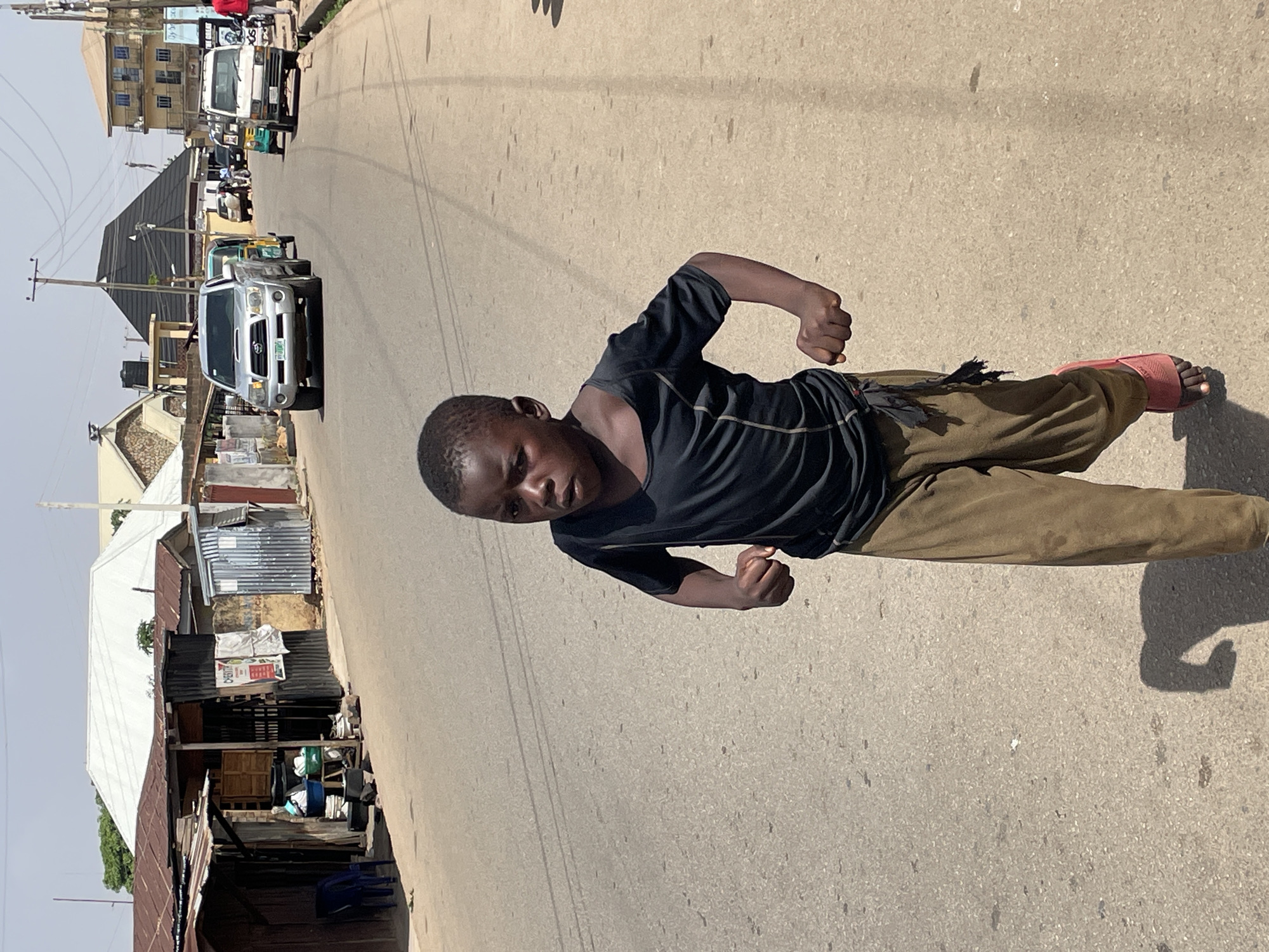 A young boy with dark skin, wearing a dark short-sleeved t-shirt and olive-green trousers, stands on a paved street. He wears red slip-on sandals. His body is angled to the right, but his head is turned, looking directly at the camera with a serious expression. Both of his hands are clenched into fists at his sides, bent at the elbows, suggesting a dynamic or playful pose.
The scene is an outdoor street illuminated by bright daylight, casting sharp shadows that indicate a clear sky and likely midday or early afternoon. In the background, the street recedes into the distance, lined by various structures and vehicles.
On the right side of the street, a silver SUV is visible, bearing a Nigerian license plate that clearly shows "JOS", consistent with the location context of Jos, Nigeria. Further along the road, a white van and a green and yellow auto-rickshaw (Keke Napep) are also present. On the left side of the street, several modest buildings with corrugated metal roofs and painted walls can be seen, including what appears to be a small, open-fronted shop or stall. Power poles and overhead wires crisscross the frame. Some indistinct text is visible on a white bag or poster on one of the background structures.