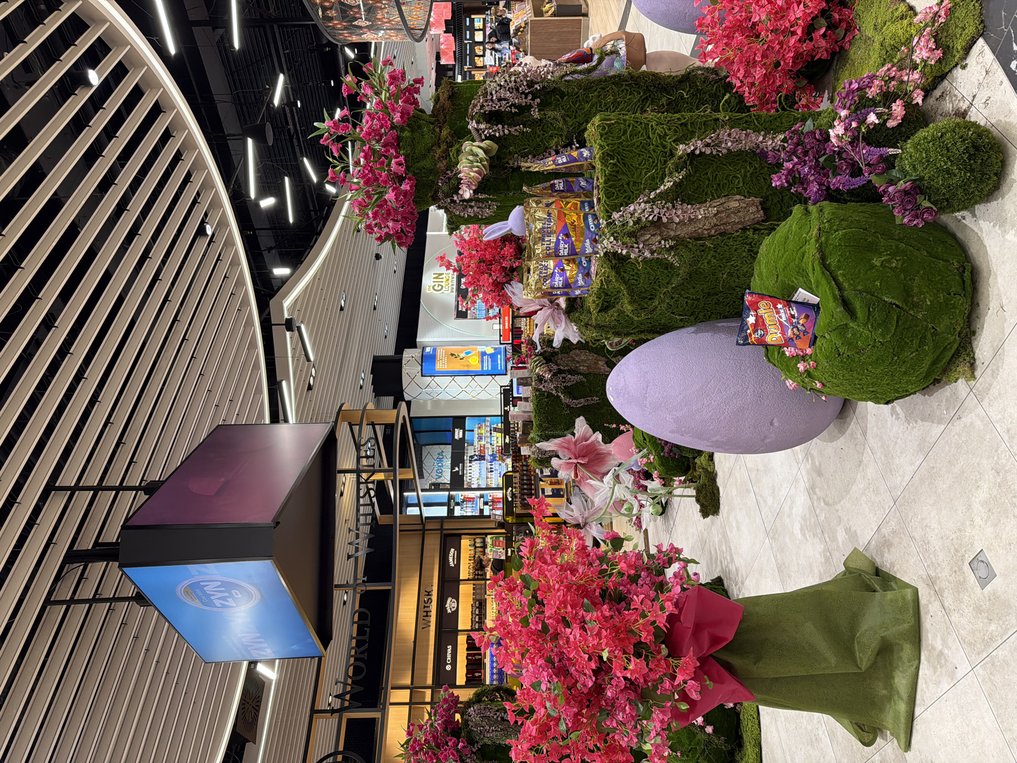 The image captures an interior space likely within Sofia Airport, Bulgaria, featuring commercial displays and decorations. The main focal point includes several large decorative features covered in green moss and adorned with pink flowers. On the display, there is confectionery, including Milka chocolate bars. The floor is tiled in a neutral tone. Behind these decorations are retail displays of alcoholic beverages, including whiskey and gin. Overhead, there is a modern architectural structure with linear white elements against a dark ceiling punctuated with lighting fixtures. There are no people visible in the close foreground, though some individuals can be seen in the background amidst the retail displays. A large hanging sign advertising alcohol brands is also visible.