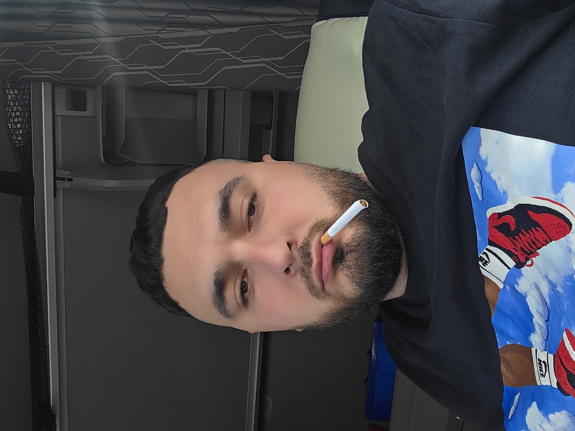 A male individual with short dark hair, a dark beard, and mustache is shown in a close-up, seated within the interior of what appears to be a vehicle cabin, specifically located in Miranda de Ebro, Spain. He is looking directly at the camera with an unlit cigarette held between his lips, the filter facing outwards. He wears a black long-sleeved shirt or sweatshirt with a graphic design on the chest area, featuring two legs adorned with red and black athletic shoes (with "NIKE" branding on white socks) against a backdrop of a blue sky and white clouds. The background behind him consists of dark grey interior panels, an overhead storage compartment with a black mesh net, and a folded grey curtain with a geometric pattern on the right. A light green cushion is visible behind his right shoulder. A blue rectangular object is partially visible on the lower left.