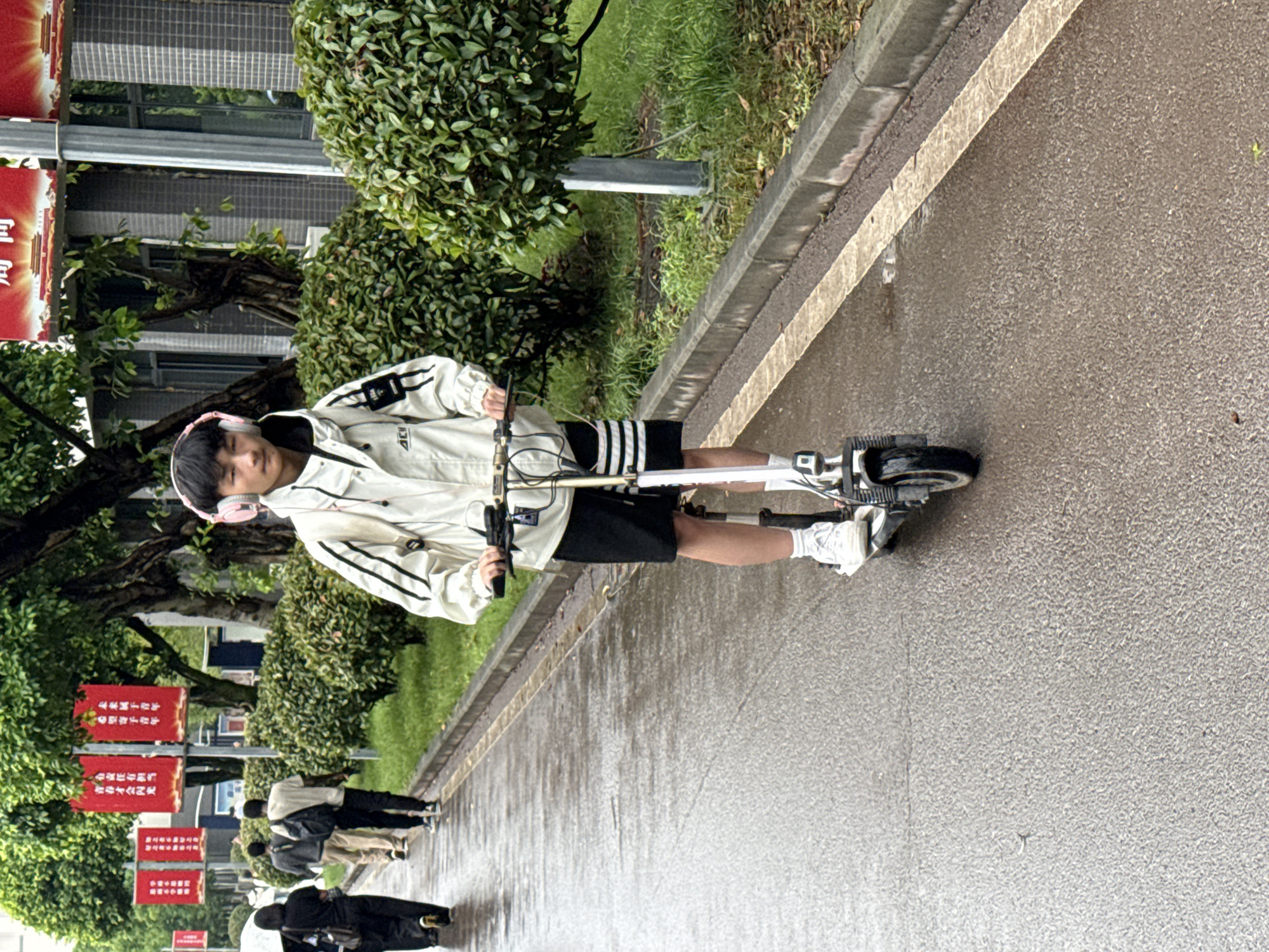 A young person is riding a scooter on a wet paved pathway, wearing a light-colored jacket with black stripes, black shorts, and white sneakers, and pink headphones. To the left are green bushes and trees. In the background, other people are walking. There are red banners with white text, likely hanging from poles. The location is Jiangjin, China.